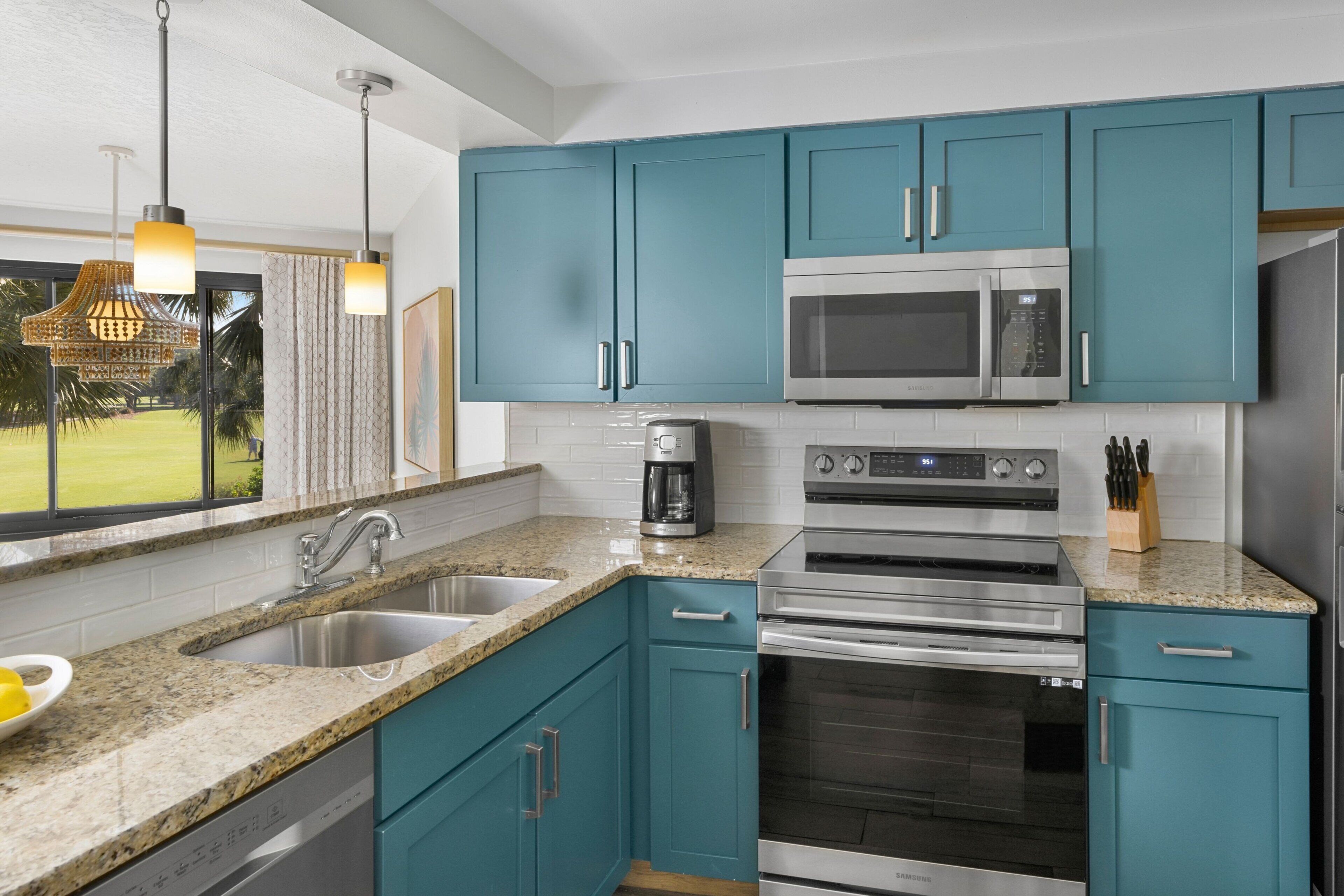 A modern kitchen with teal cabinets, stainless steel appliances, granite countertops, and a large window offering a view of a green lawn.