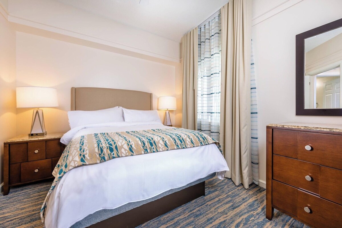 Habitación de hotel con una cama, dos mesitas de noche con lámparas, un aparador y un espejo grande en la pared.