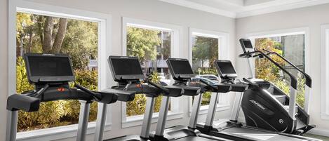 A row of treadmills with digital displays, large windows with a view of trees and a parking lot, and a clean, modern interior.