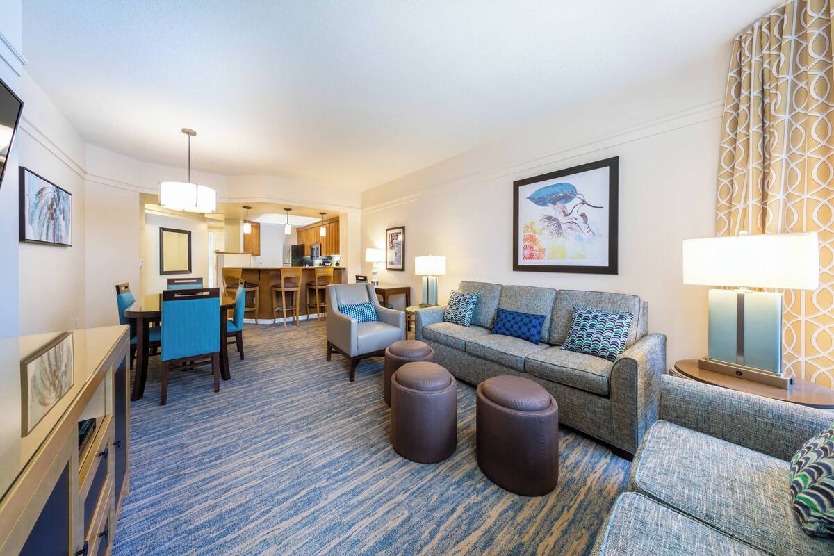 Habitación de hotel con sala de estar que incluye un sofá, otomanes y un comedor. Se observa una cocina al fondo.