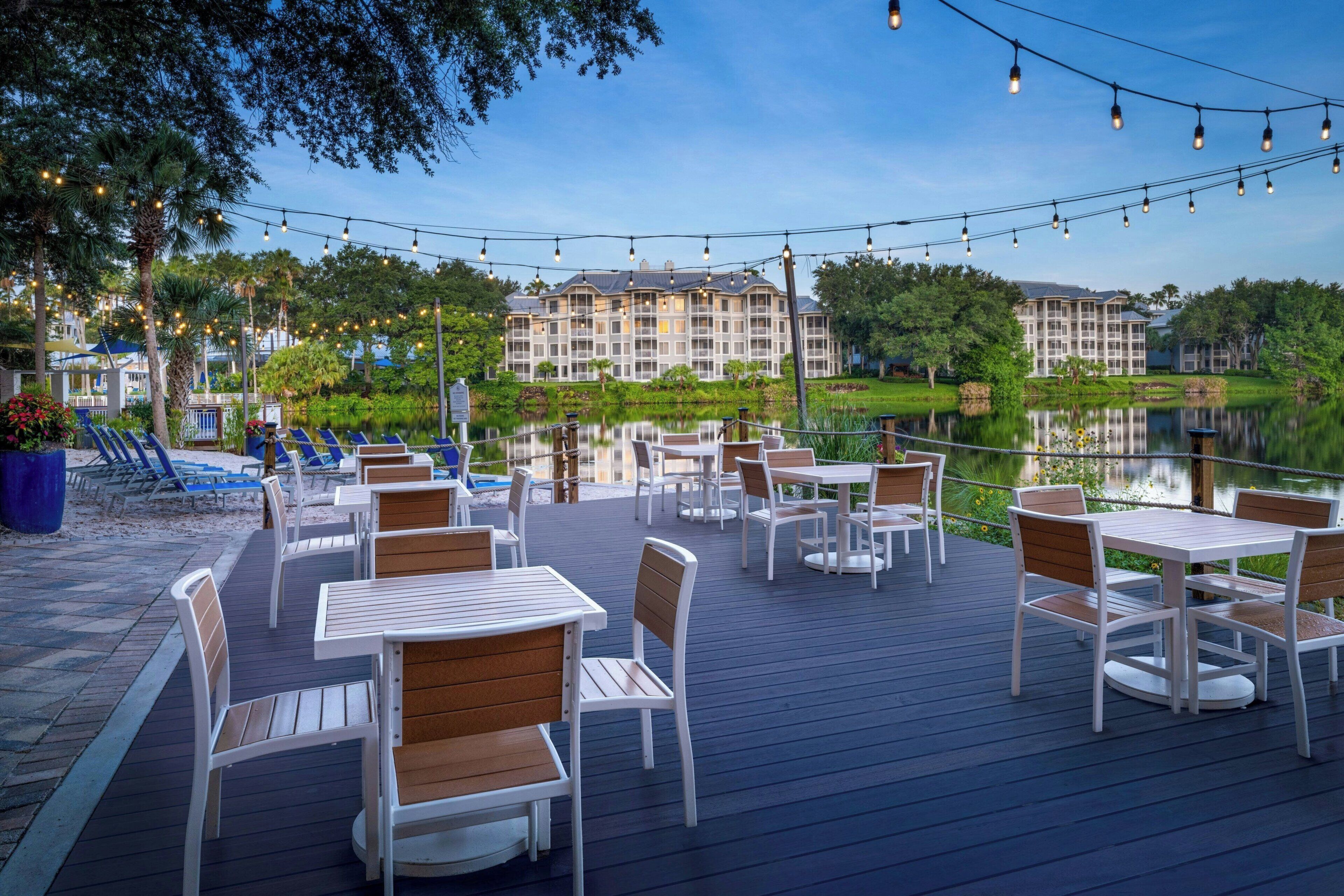 An outdoor deck with tables and chairs, overlooking a body of water and a building with multiple balconies.