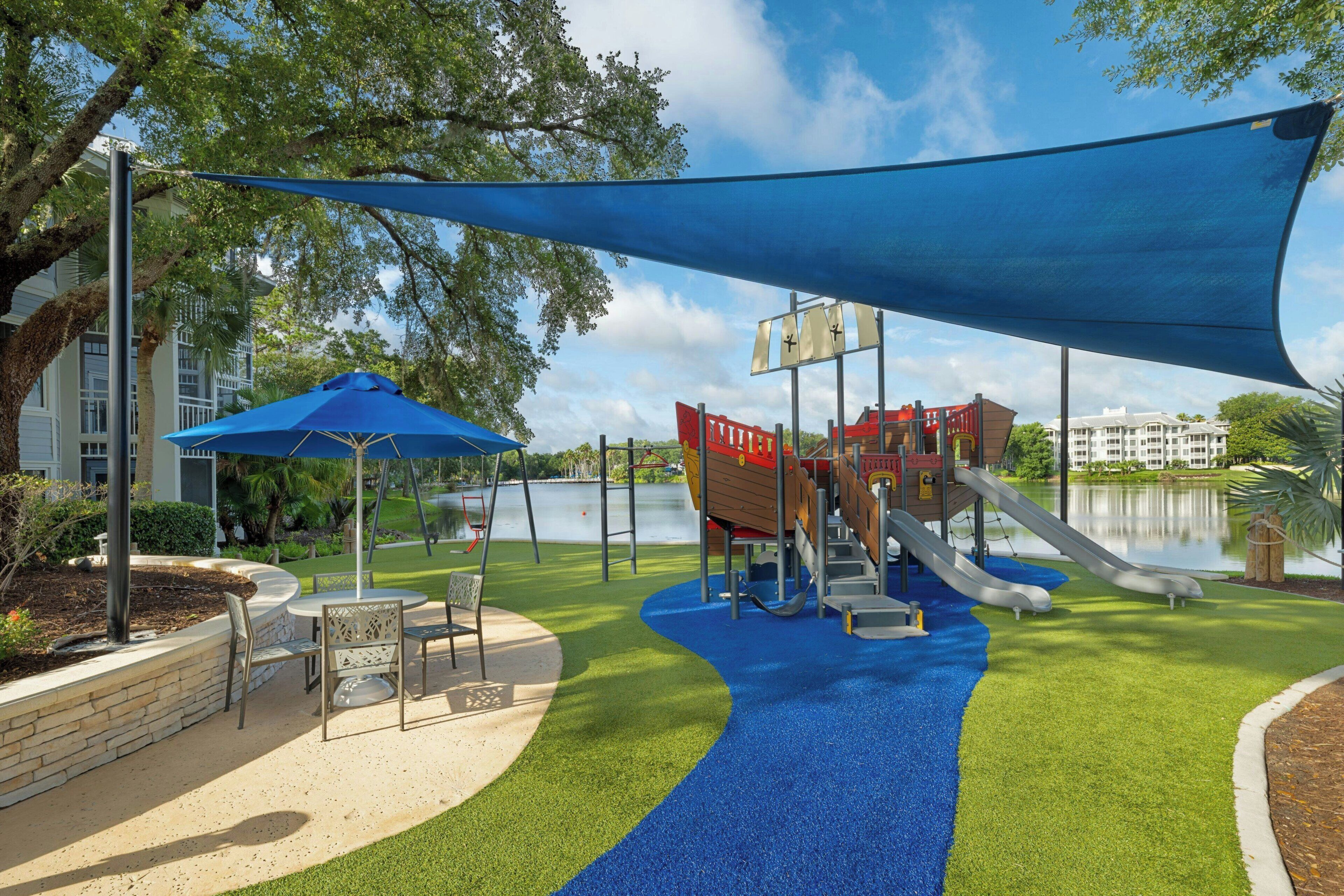 An outdoor playground with slides and climbing structures, surrounded by a lake and trees.