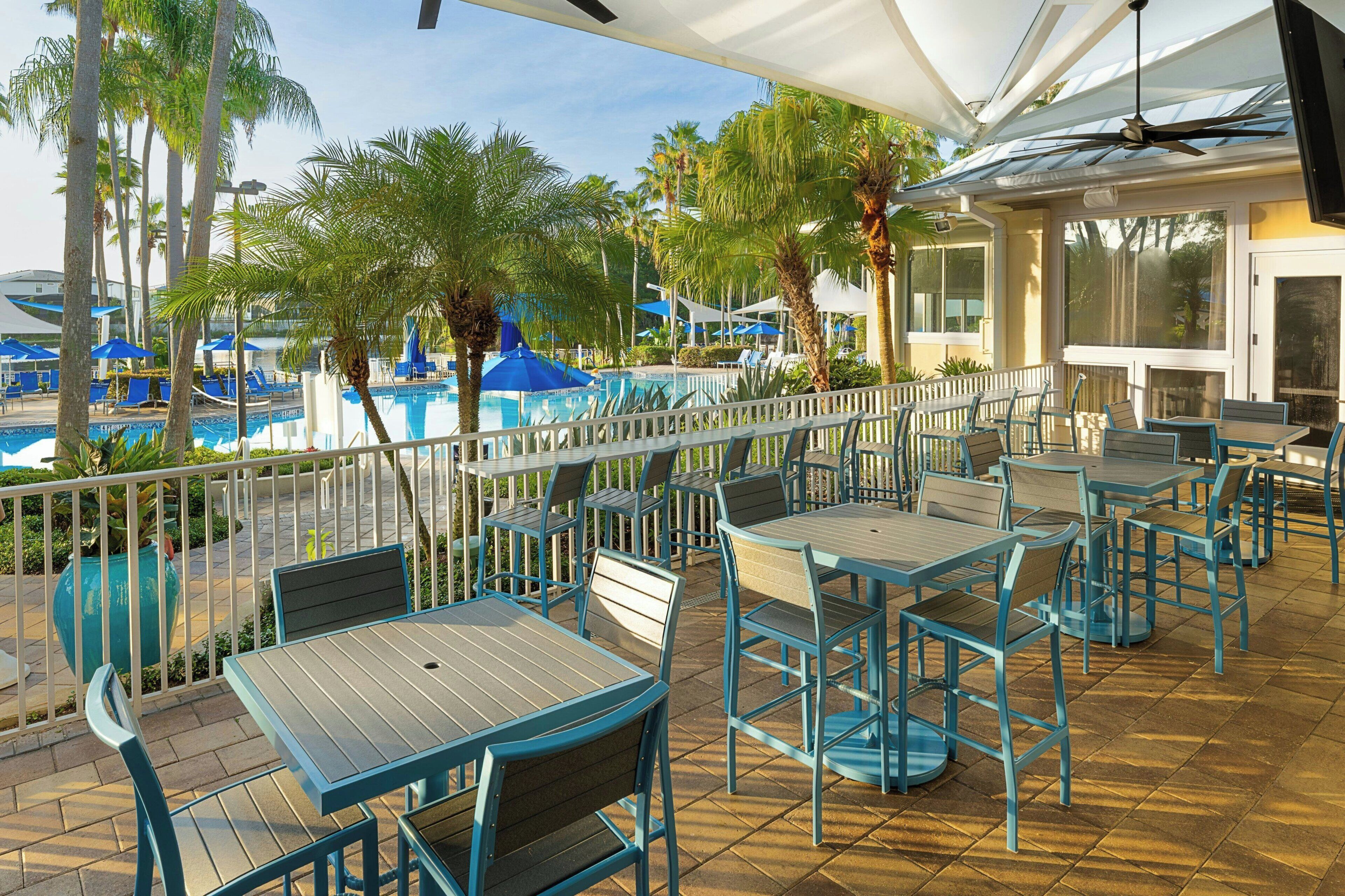Un patio al aire libre con mesas y sillas, con vista a una piscina y palmeras.