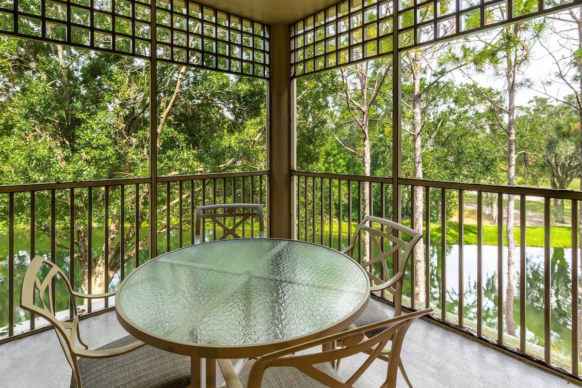A glass-top table with chairs on a balcony with metal railings, overlooking a body of water and trees.