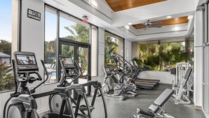 Un gimnasio con diversos equipos de ejercicio, ventanas amplias y un ventilador de techo.