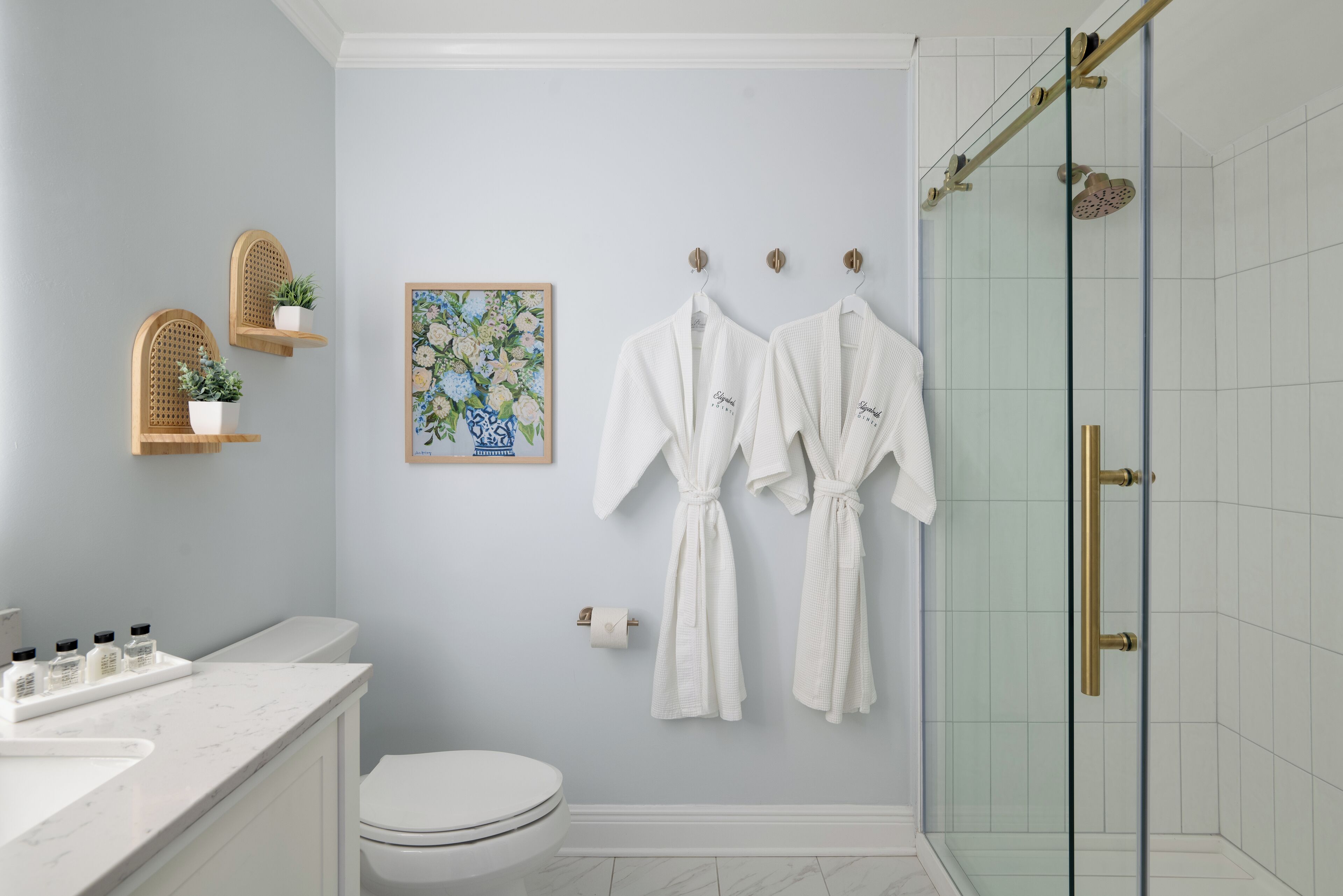 A bathroom with a toilet, a sink with toiletries, a shower with glass doors, and two white robes hanging on hooks.
