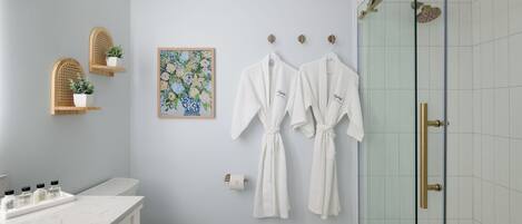 Un baño con inodoro, lavamanos con artículos de aseo, ducha con puertas de vidrio y dos albornoces blancos colgados en ganchos.