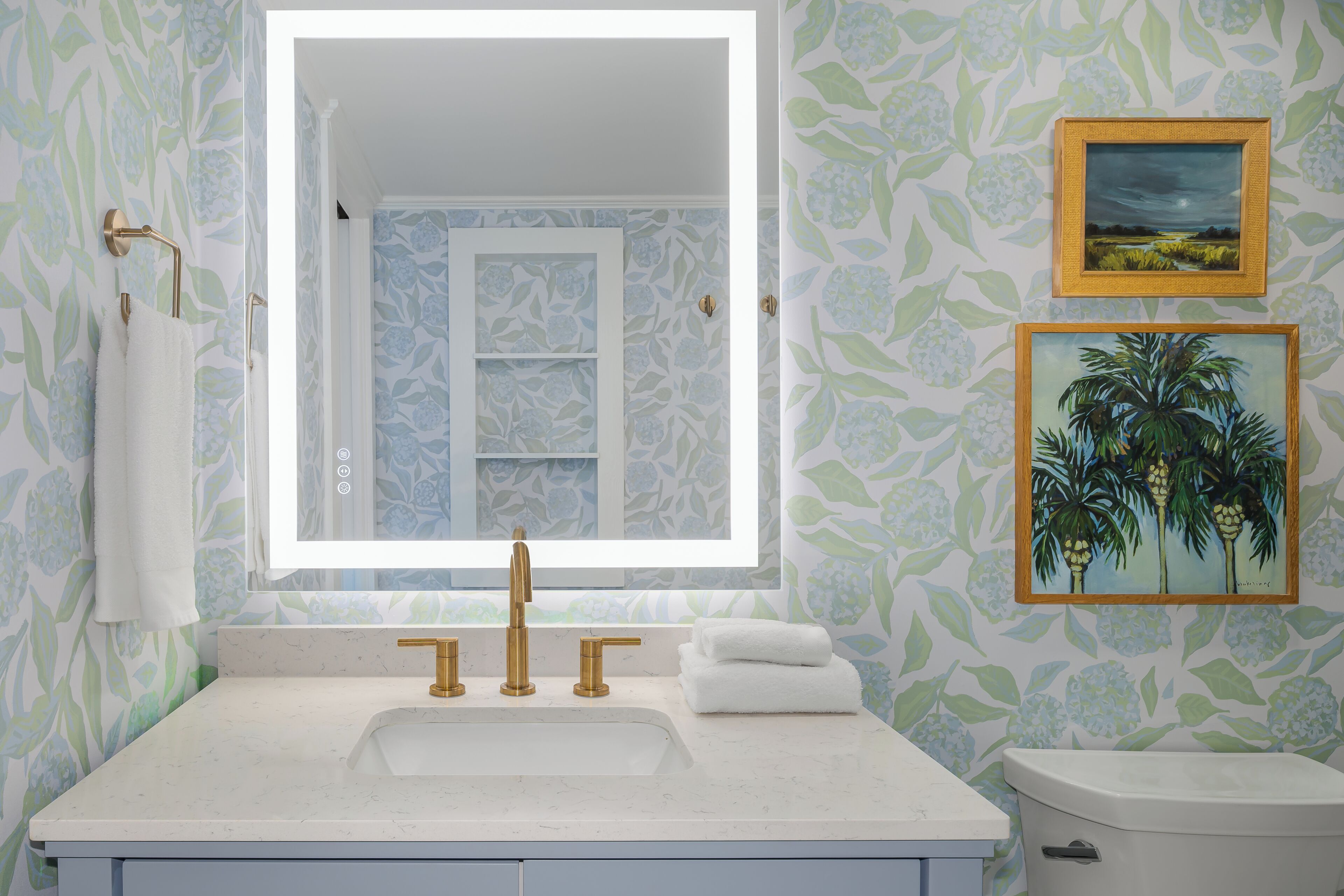 A bathroom with a large mirror, a white sink, and two gold faucets. There are two framed pictures on the wall, one of a landscape and one of palm trees.