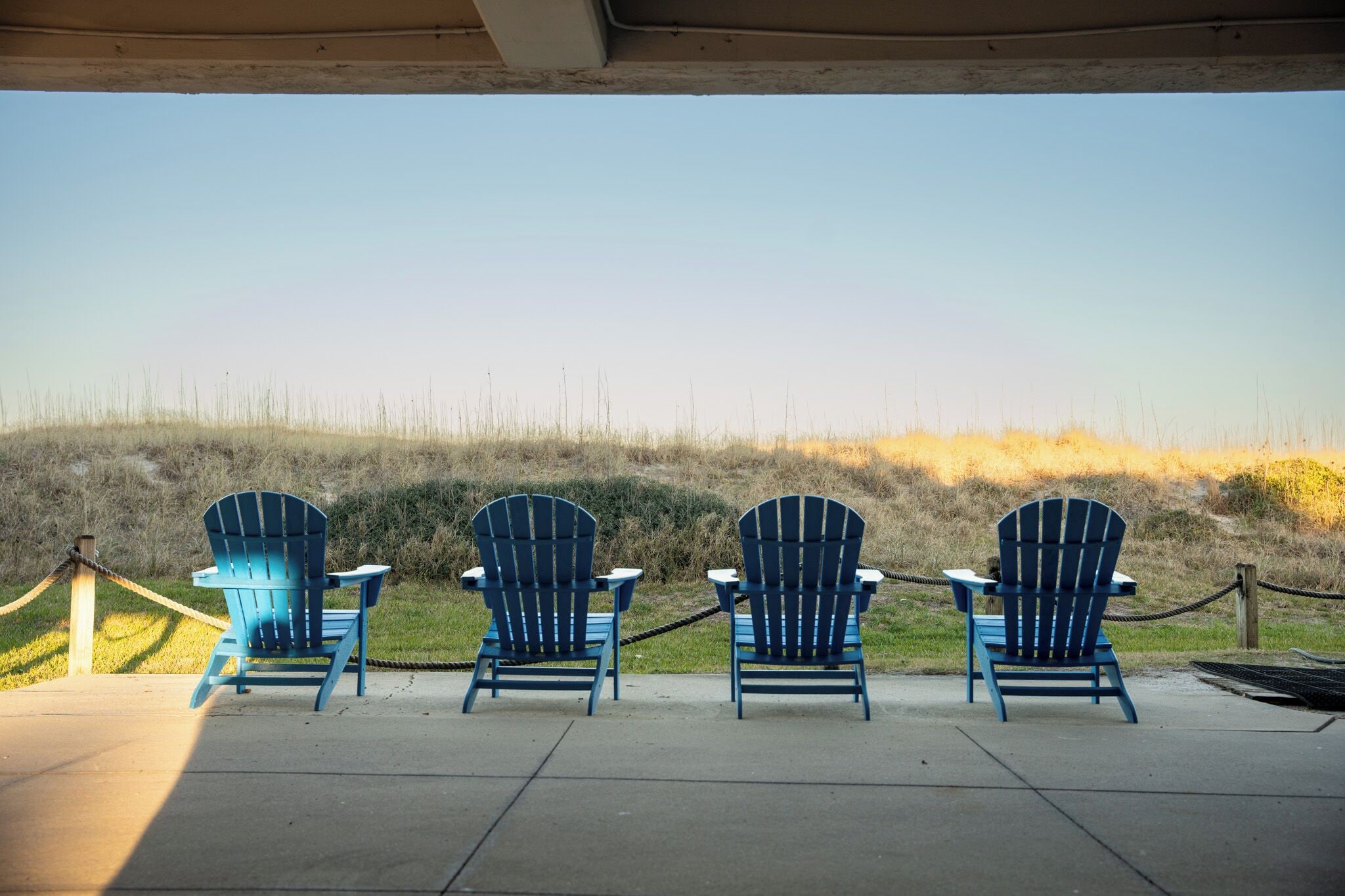 Four blue Adirondack chairs lined up on a concrete surface with a view of a grassy hill in the background.