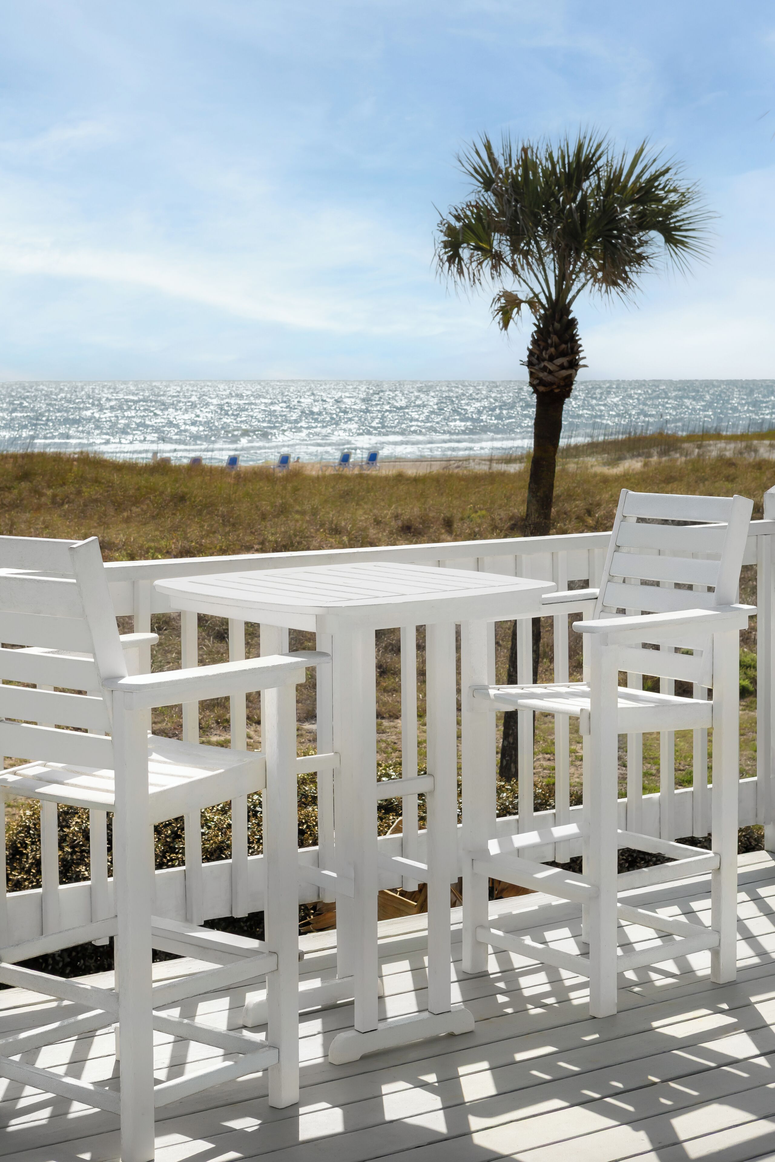 Un espace extérieur aménagé avec des meubles blancs, comprenant une table et des chaises, offrant une vue sur une plage, un palmier et l’océan.
