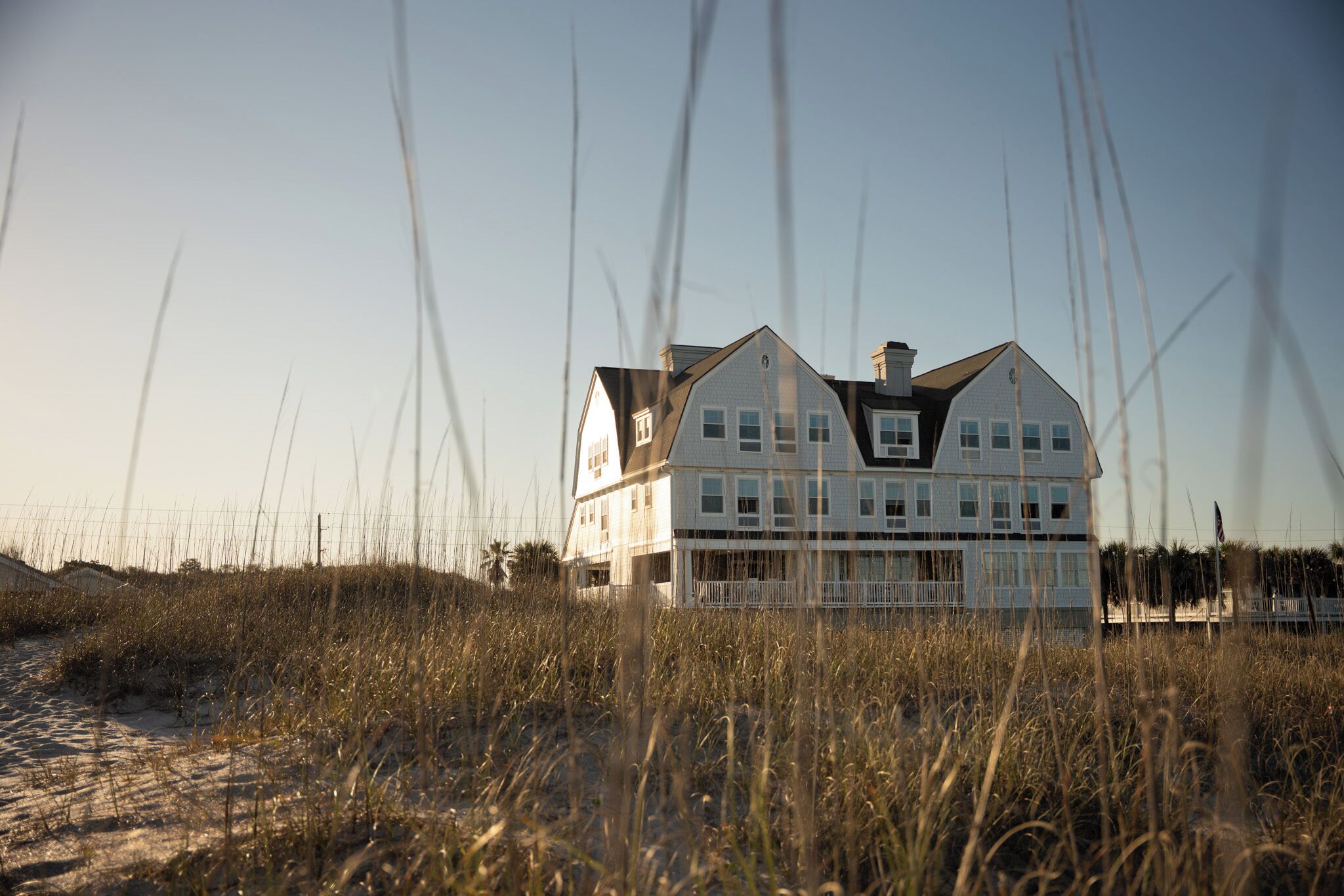 A large, multi-story white building with a symmetrical roofline, situated on a sandy beach with tall grass in the foreground.