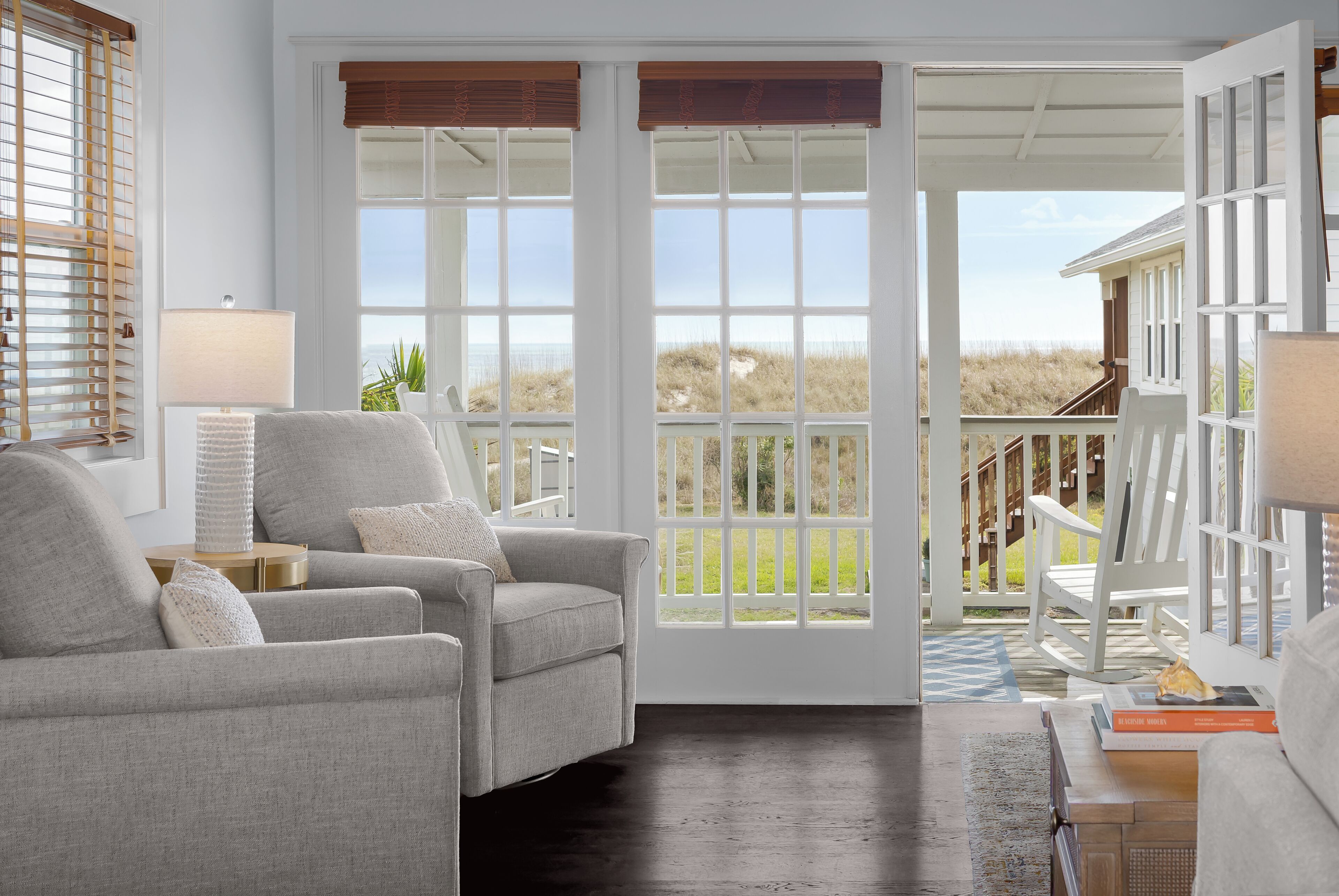A room with a grey sofa, a lamp, and a view of the beach through large windows.