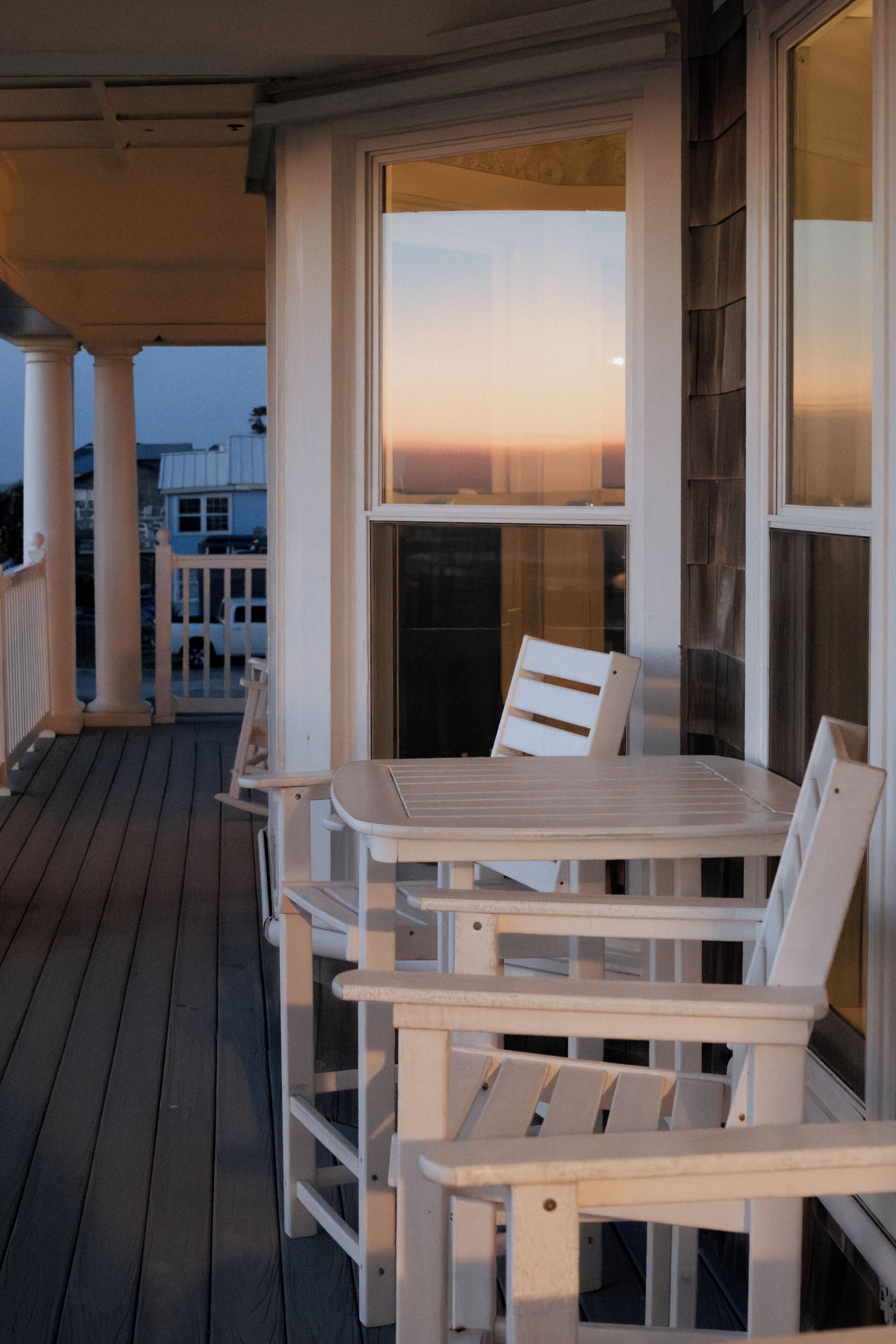 A wooden deck with a table and chairs, a railing, and a view of the sunset through a window.