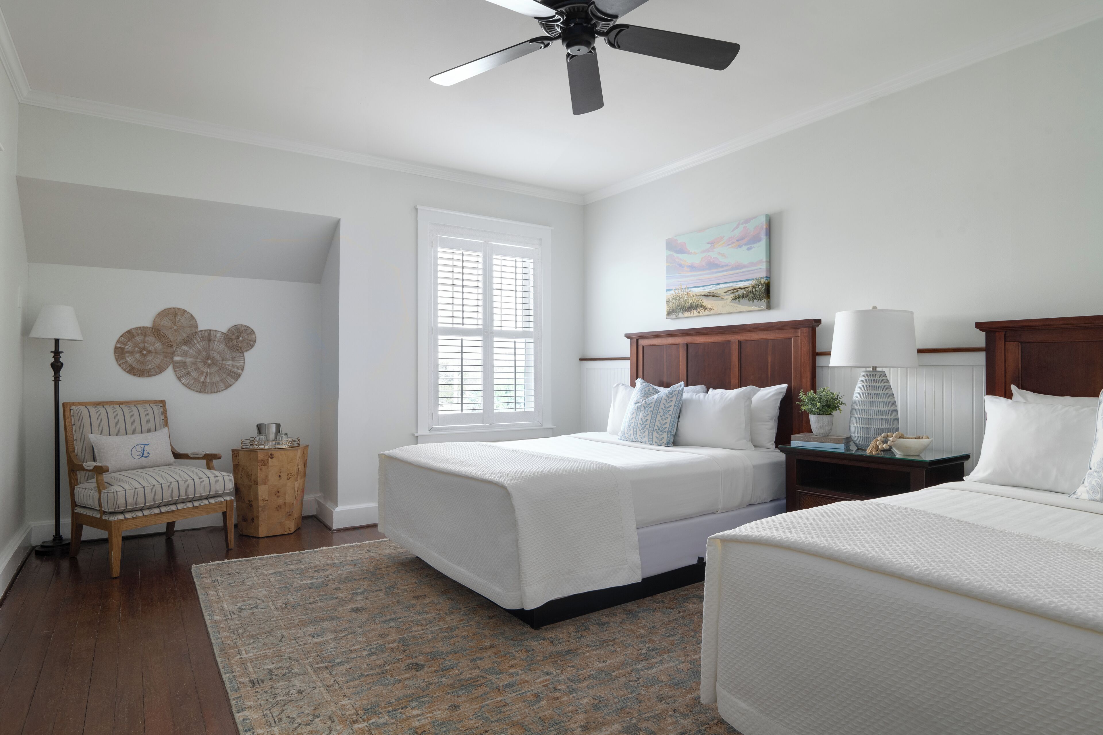 A bedroom with two beds, a ceiling fan, a window with blinds, a chair, a side table, and a painting on the wall.