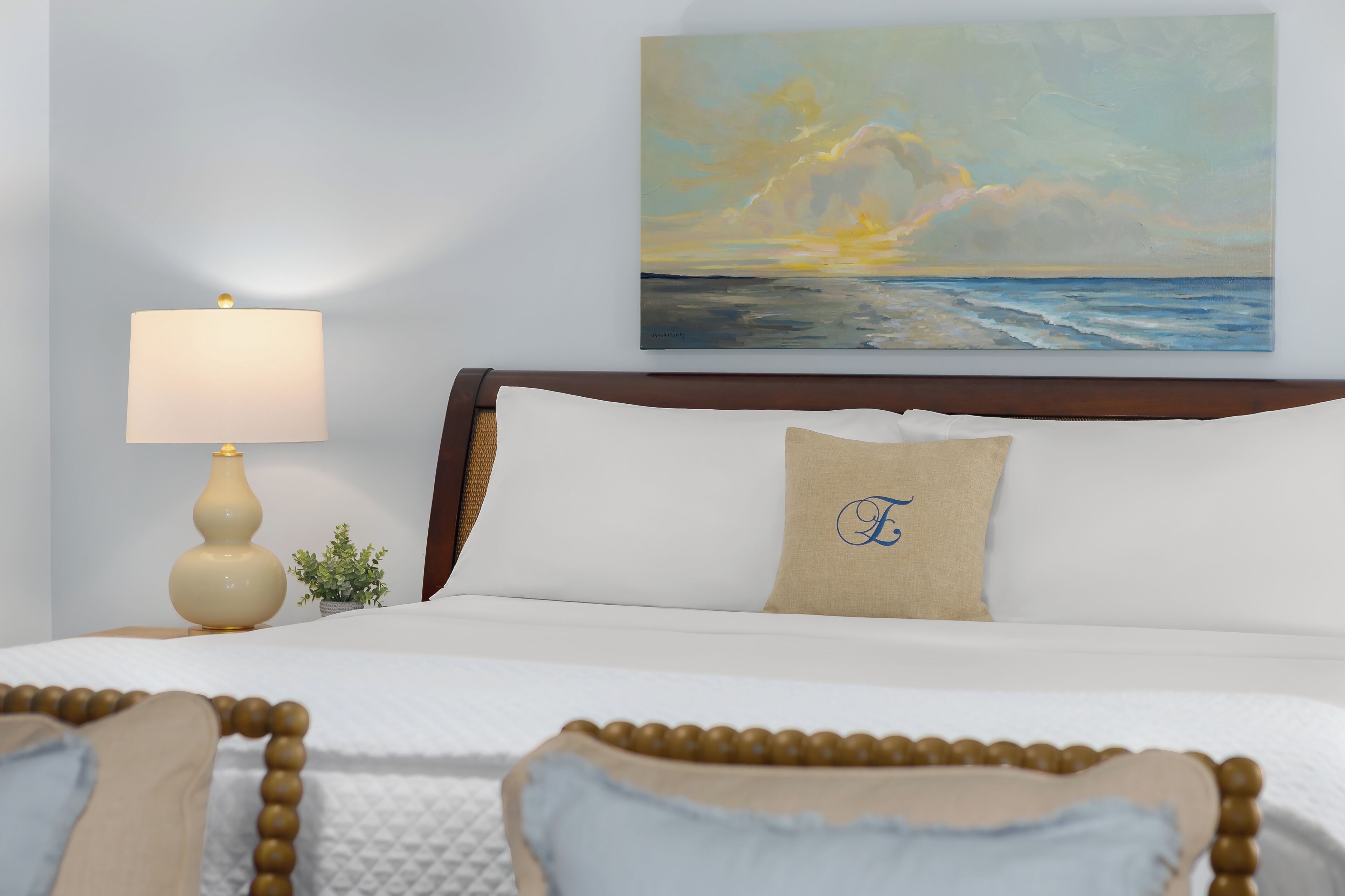 A neatly made bed with a headboard, a bedside lamp, a painting of a sunset over the ocean, and a pillow with a monogram.