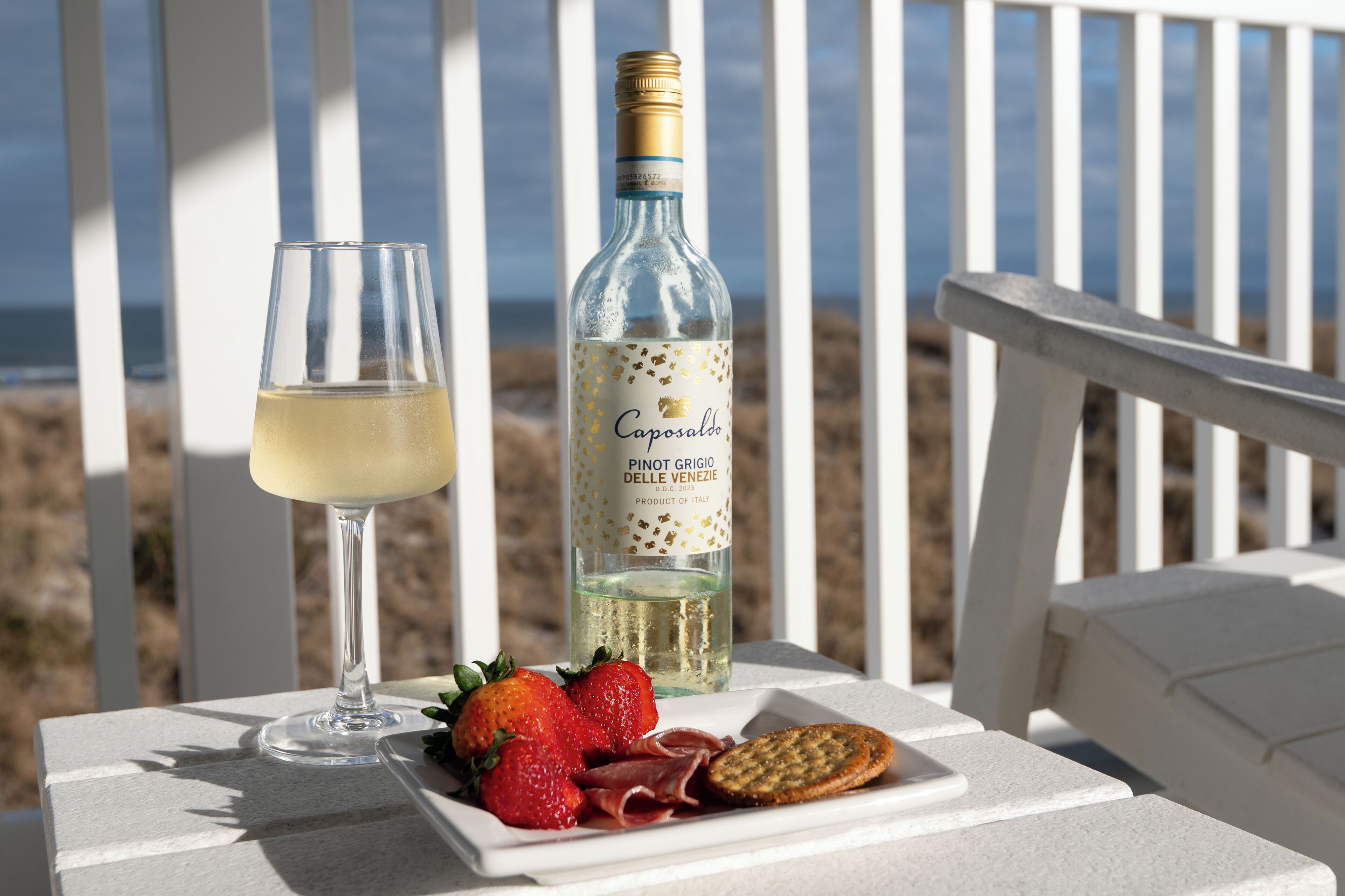 Una botella de Caposaldo Pinot Grigio, una copa de vino blanco, fresas y una rebanada de jamón curado sobre una mesa blanca con vista a la playa.