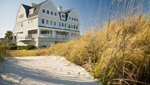 Een groot, twee verdiepingen tellend huis met een balkon, gelegen nabij een zandstrand met hoge grassen.