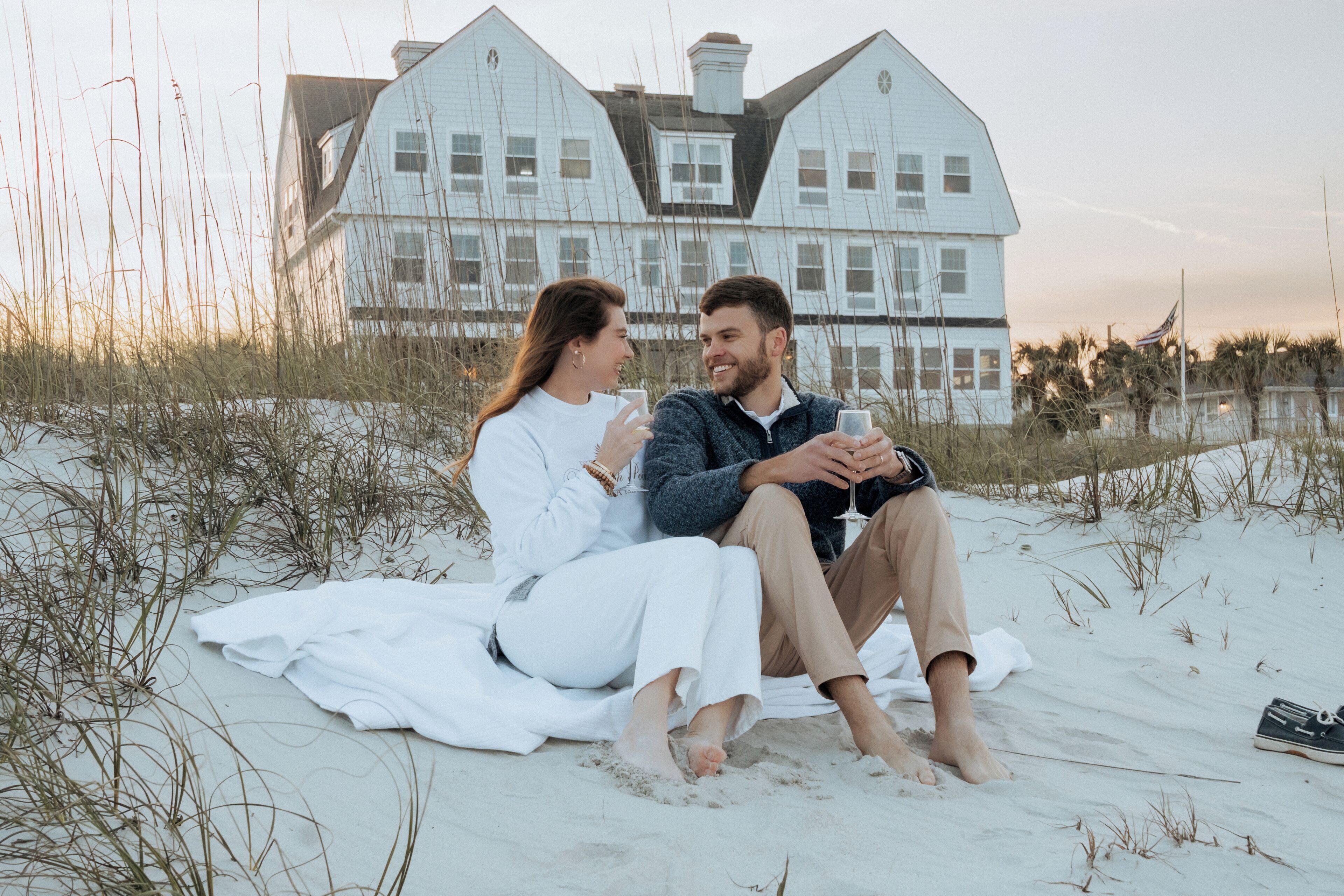Two people sitting on a beach with a house in the background.