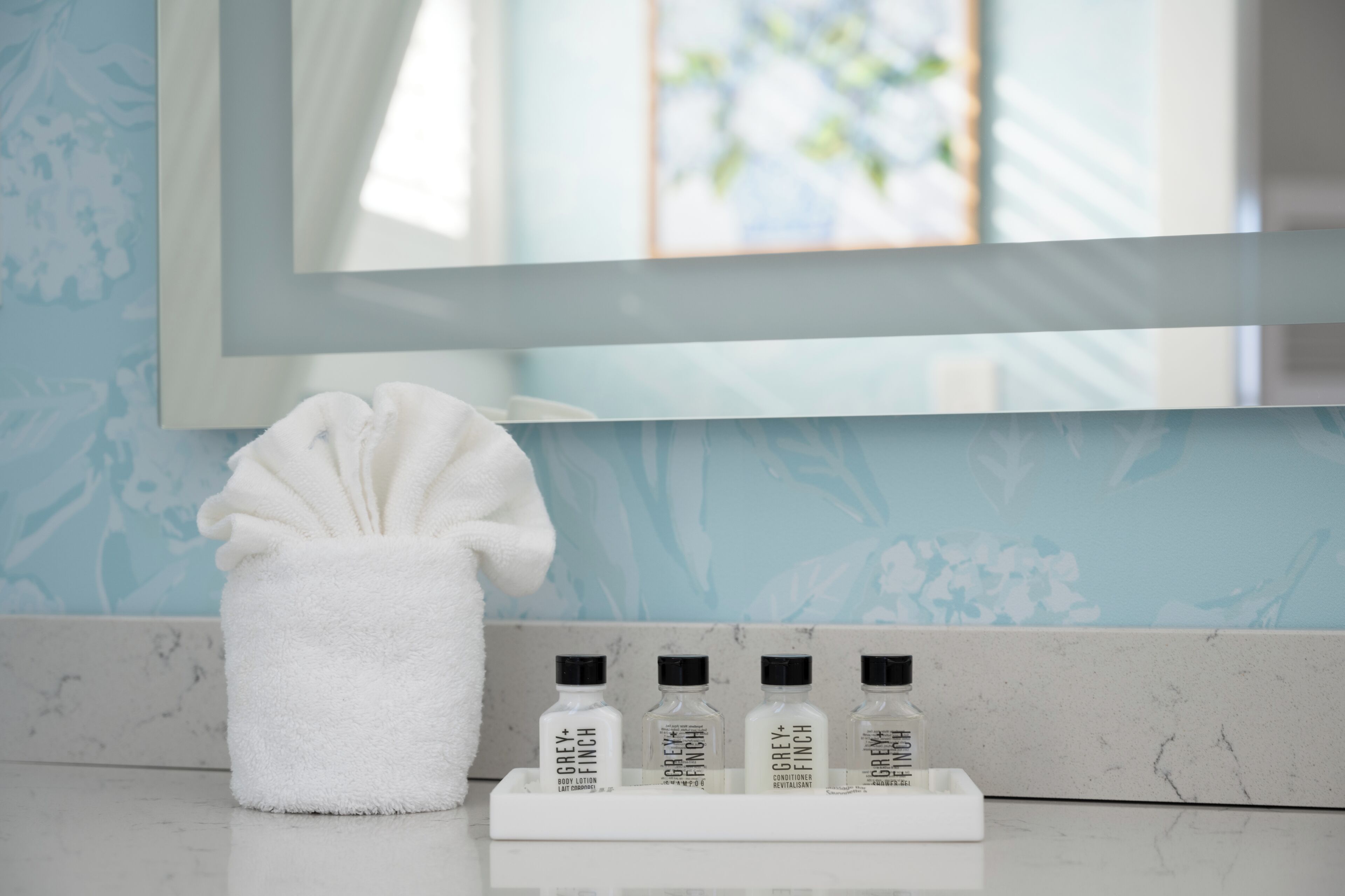 Une salle de bain avec une serviette blanche, un miroir et trois flacons de lotion pour le corps de la marque Grey Finch, disposés sur un plateau.