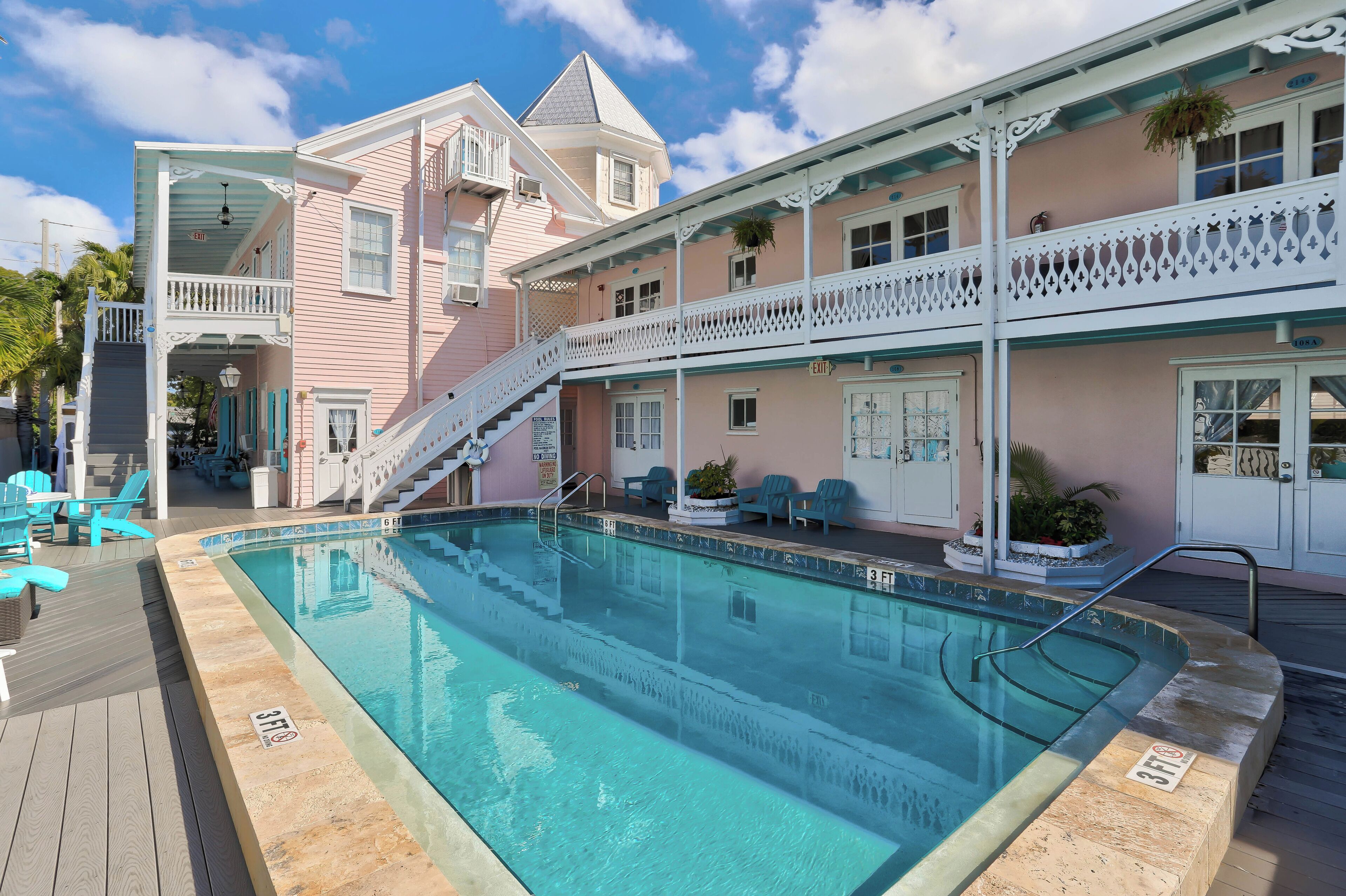 Zona de piscina con una escalera que conduce a un balcón, rodeada por un edificio rosa con barandillas blancas y un cartel que indica una profundidad de 3 pies.