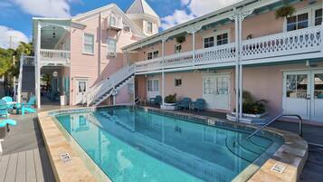 Zona de piscina con una escalera que conduce a un balcón, rodeada por un edificio rosa con barandillas blancas y un cartel que indica una profundidad de 3 pies.