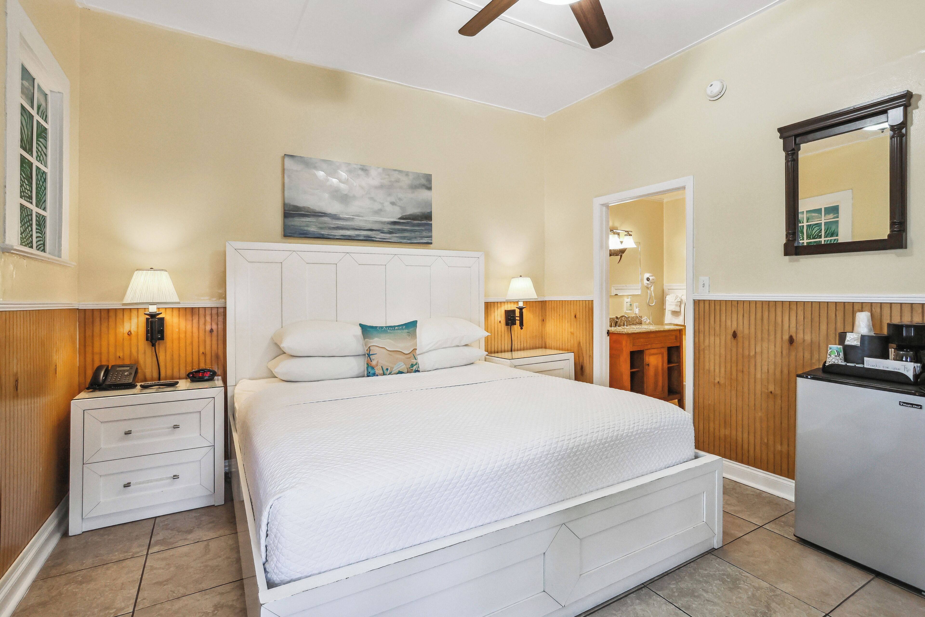 Une chambre à coucher comprenant un lit, des tables de chevet, une armoire en bois, un réfrigérateur et un ventilateur de plafond.