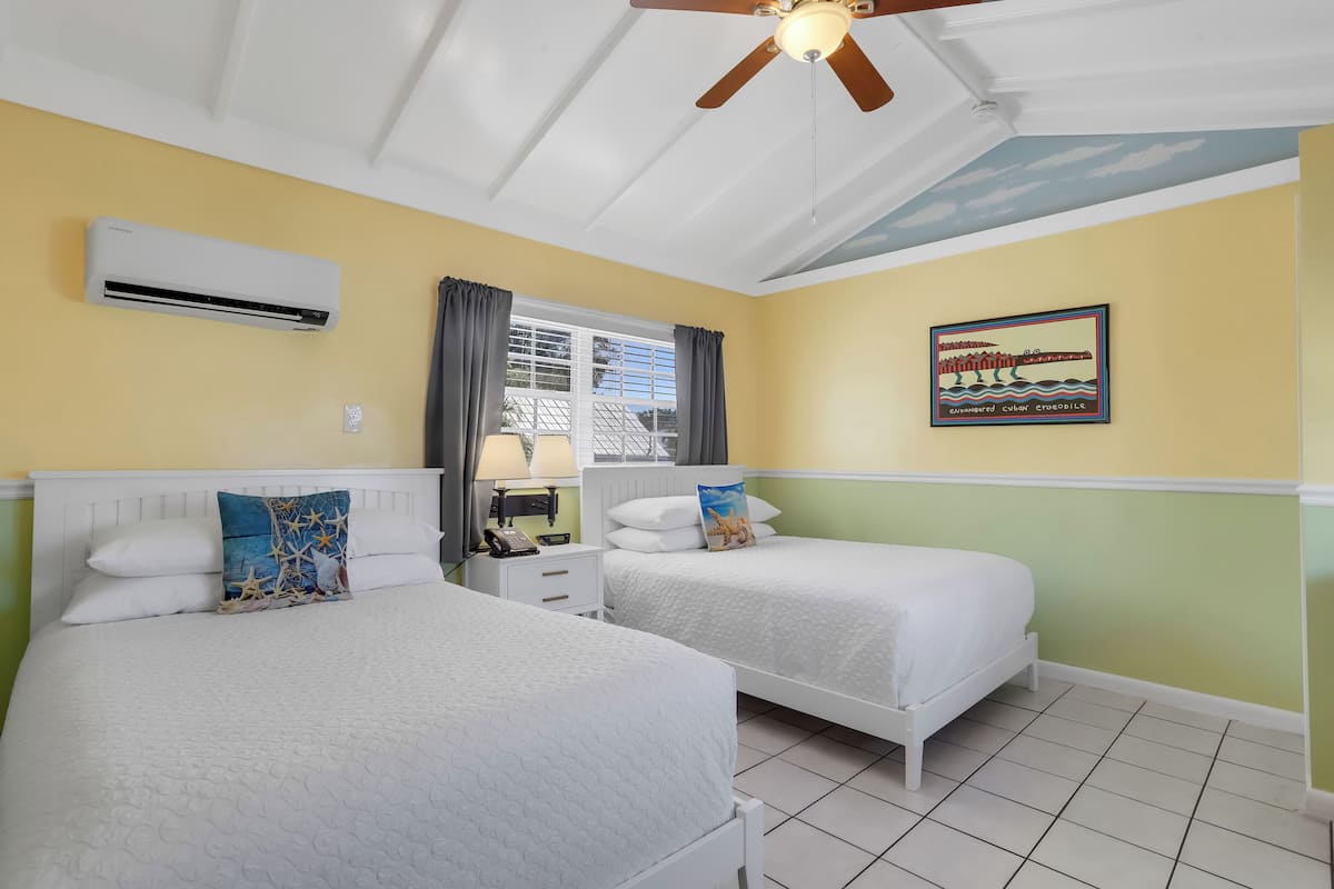 Une chambre d’hôtel avec deux lits, un ventilateur de plafond, un appareil de climatisation, une fenêtre avec des rideaux, une table de chevet avec une lampe et un tableau au mur.