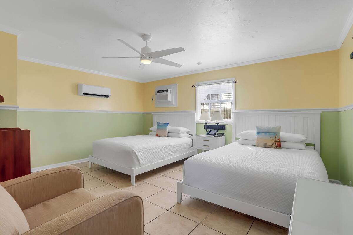 Une chambre d’hôtel avec deux lits simples, un ventilateur de plafond, un appareil de climatisation, une fenêtre avec des stores et une petite table de chevet avec une lampe.