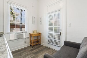 A room with a gray sofa, a wooden side table with a lamp, and a door with blinds.