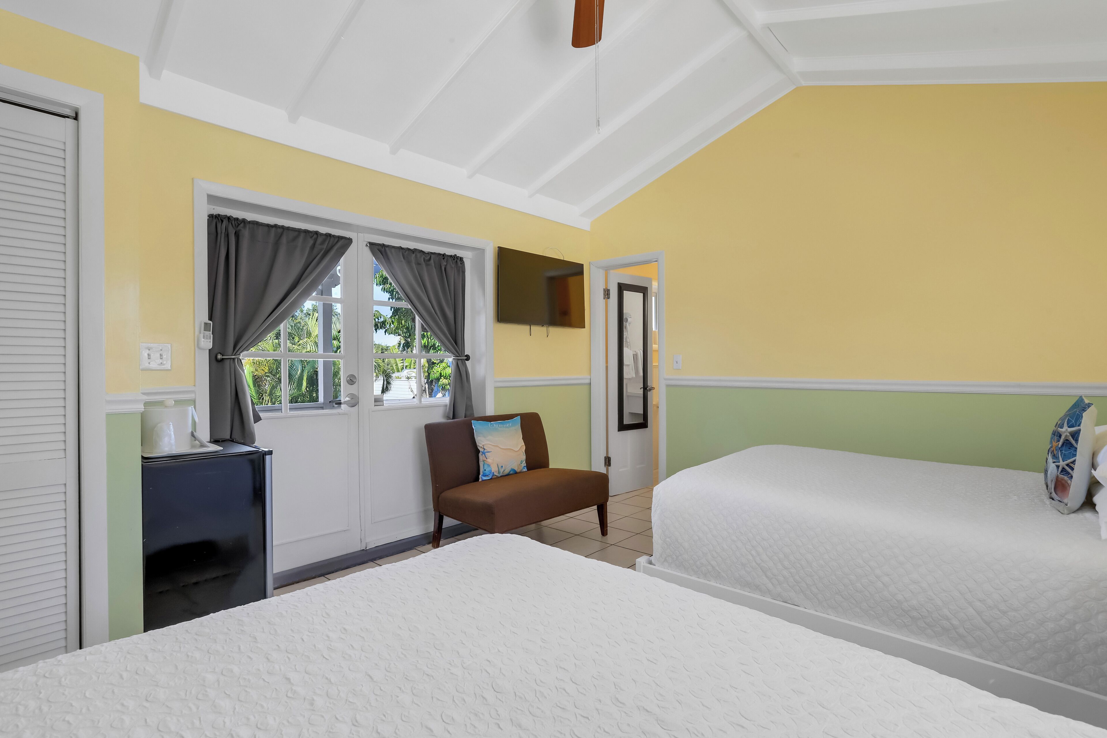 A room with two beds, a small table, a chair, a window with curtains, and a door.