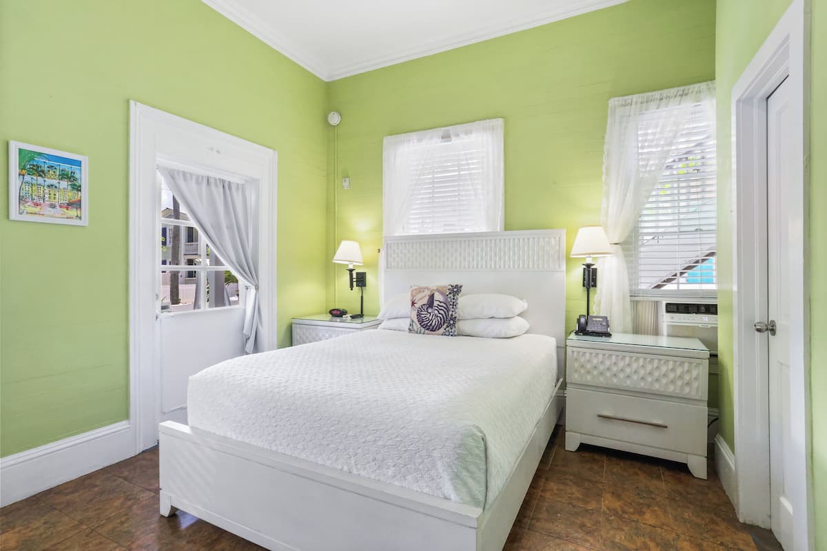 Une chambre à coucher comprenant un lit, deux tables de chevet, une commode, une fenêtre avec des stores et une porte.
