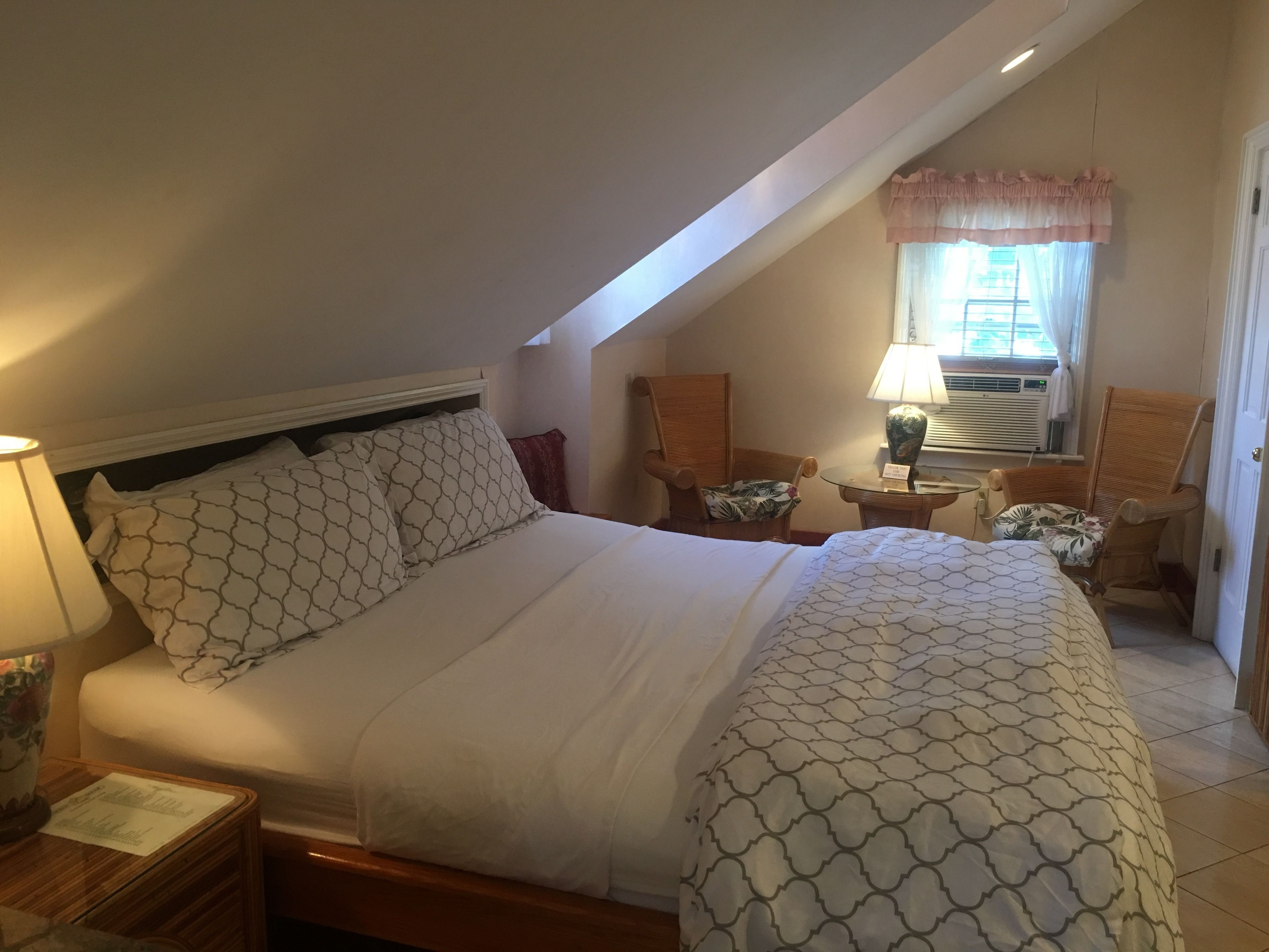 A neatly made bed with patterned bedding, a bedside table with a lamp, and a chair with a floral cushion.
