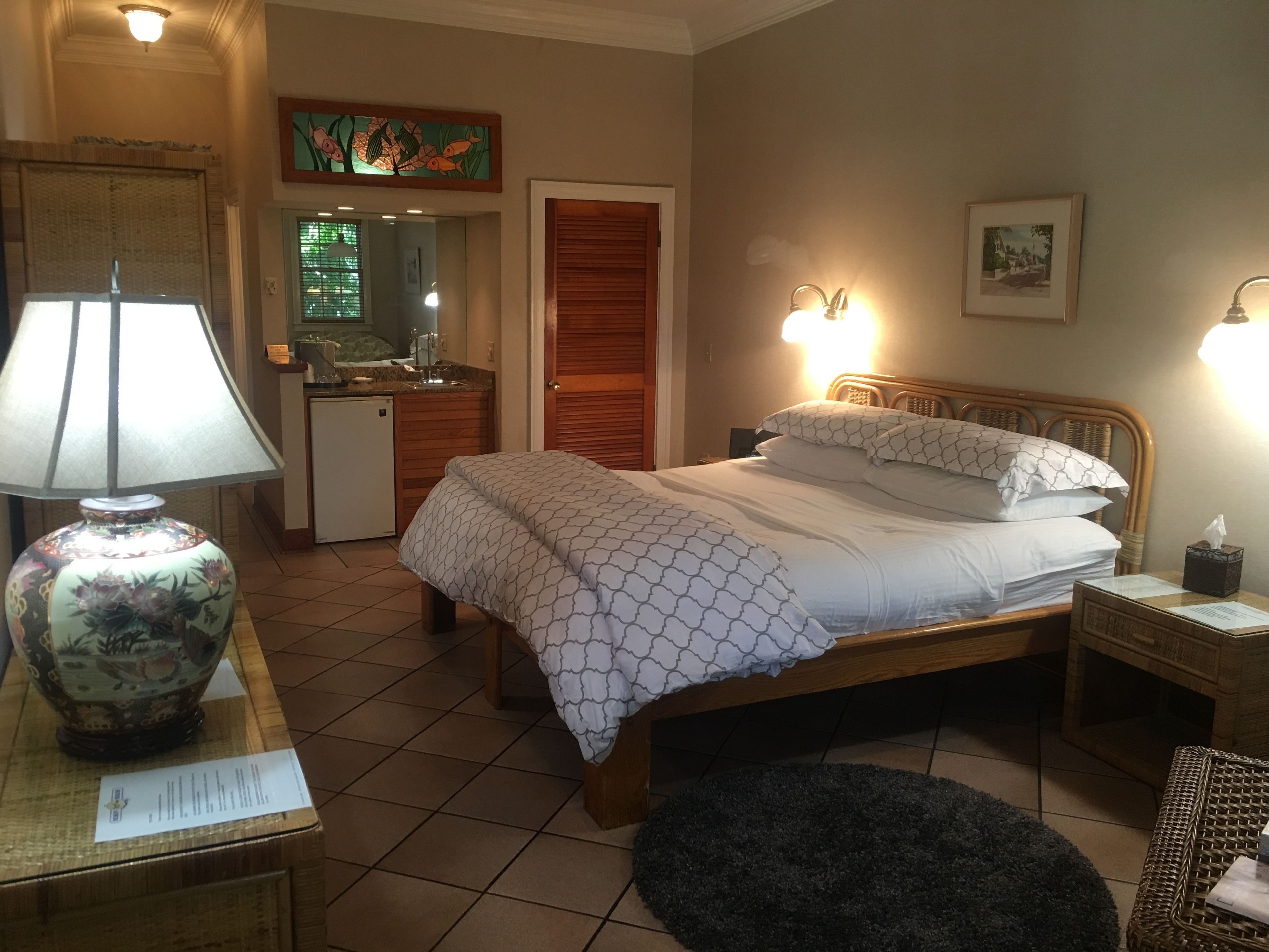 Habitación de hotel con cama, lámparas de noche, un escritorio con lámpara, un escritorio pequeño, un espejo, un cuadro en la pared y una ventana con persianas.