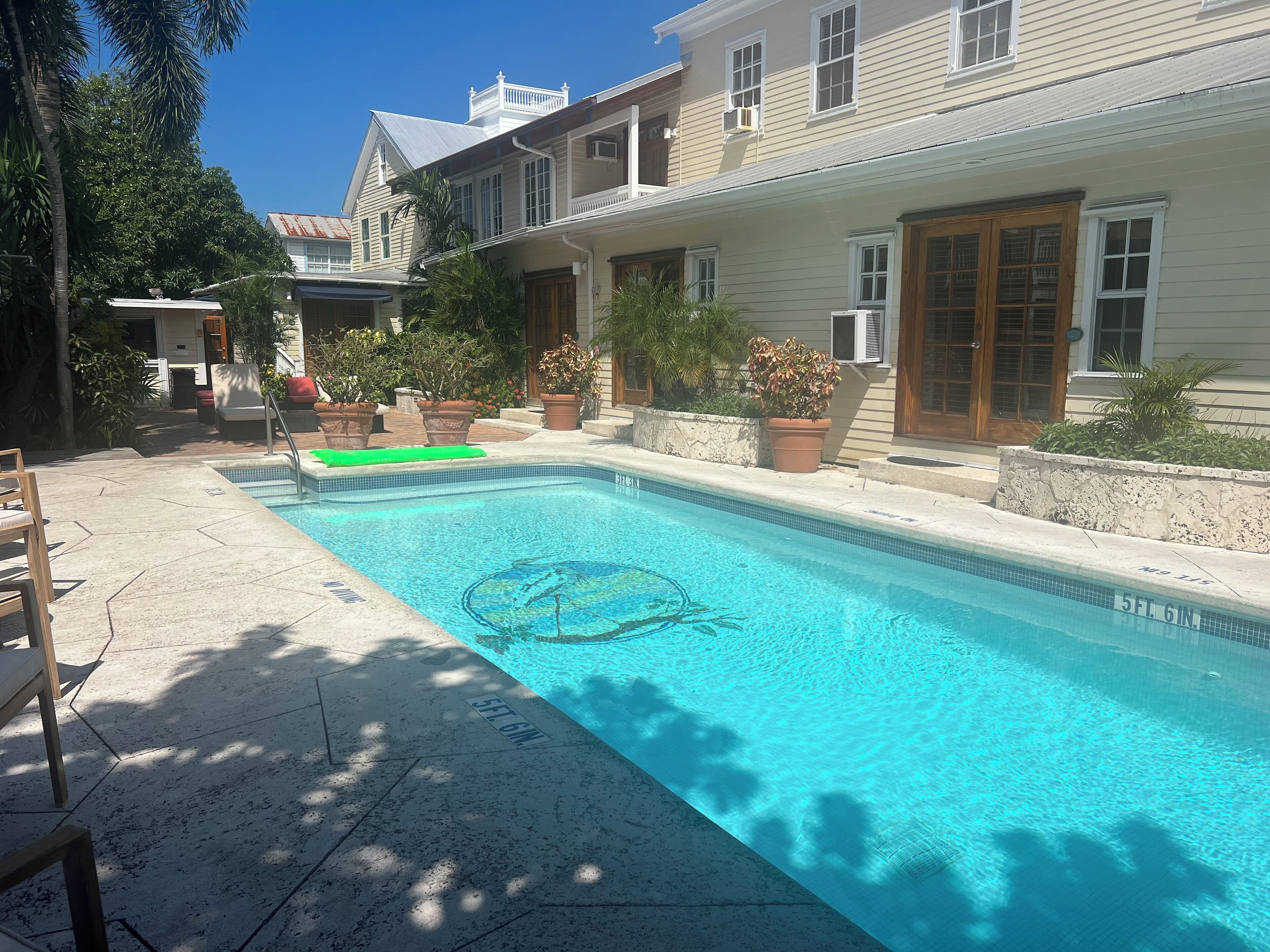 Una piscina rectangular con agua azul cristalina, rodeada por una terraza de piedra. Se ven plantas en macetas y una silla de madera.