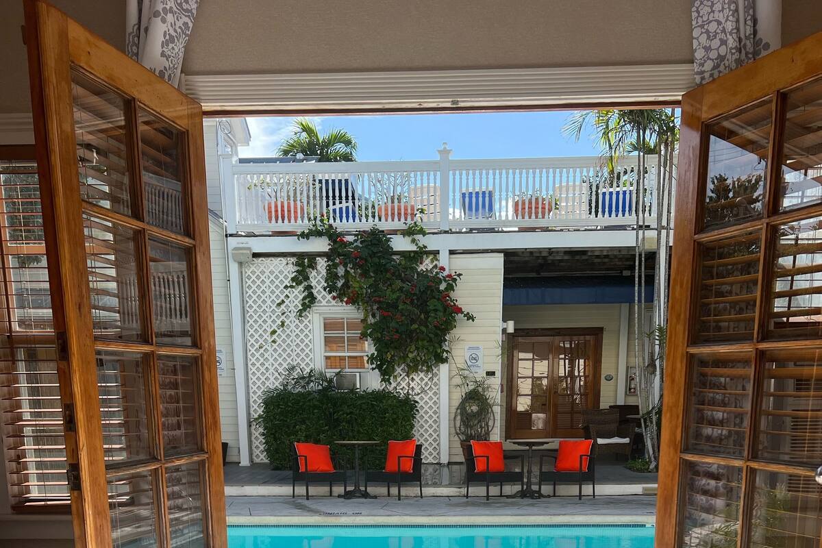 Una habitación de hotel con una cama grande, vista a una piscina y un balcón con mobiliario de exterior.