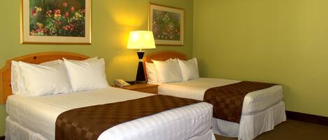 A hotel room with two beds, a nightstand with a lamp, and two framed floral paintings on the wall.