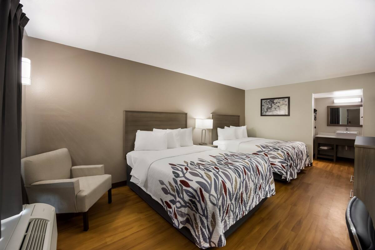 A hotel room with two beds, a desk, a chair, a lamp, and a bathroom with a sink and mirror.
