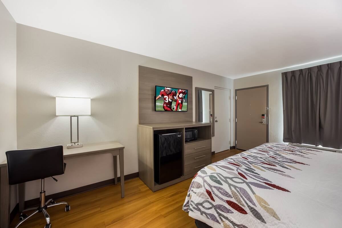 A hotel room with a bed, desk, chair, TV, and a small kitchenette.