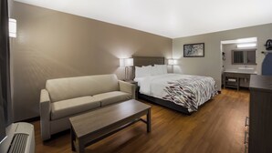 A hotel room with a bed, a sofa, a small table, a desk with a mirror, and a bathroom with a sink and toilet. - Red Roof Inn & Suites Thomasville (Thomasville)