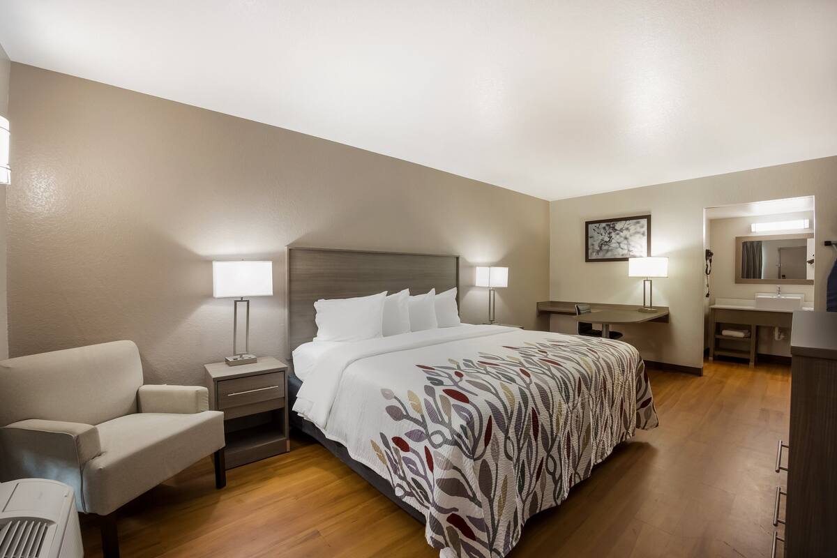 A hotel room with a bed, a desk, a chair, a nightstand, a lamp, and a bathroom.