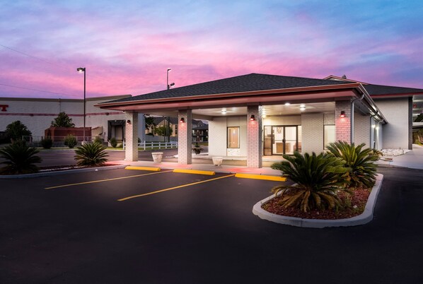 Un motel con área de estacionamiento techada, fachada de ladrillo y entrada ajardinada.