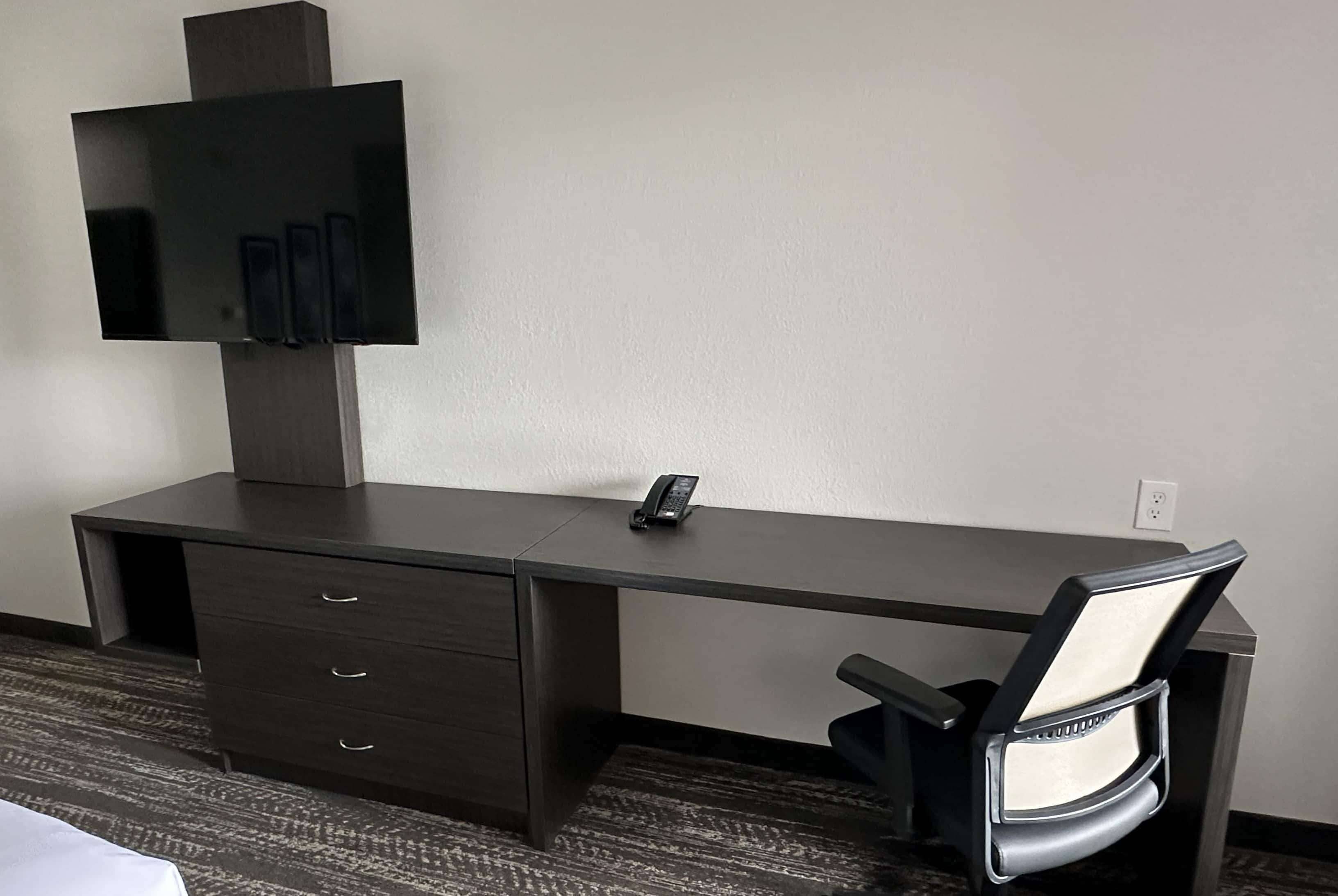 A hotel room with a dark wooden desk, a flat-screen TV mounted above it, a telephone on the desk, and a chair with a cushioned backrest.