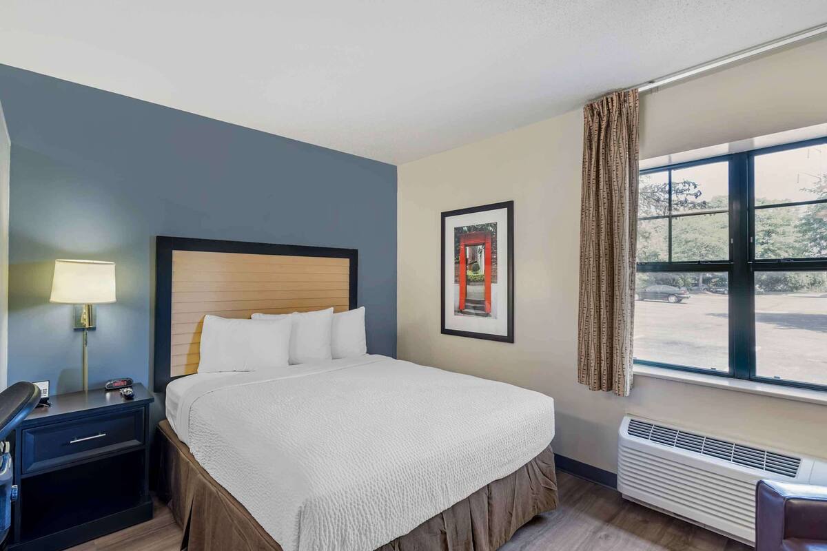 A hotel room with a bed, nightstand, lamp, framed picture, window with curtains, and a view of the outside.