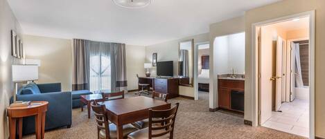 A hotel room with a dining area, a sofa, a TV, a desk, and a bathroom visible through an open door.