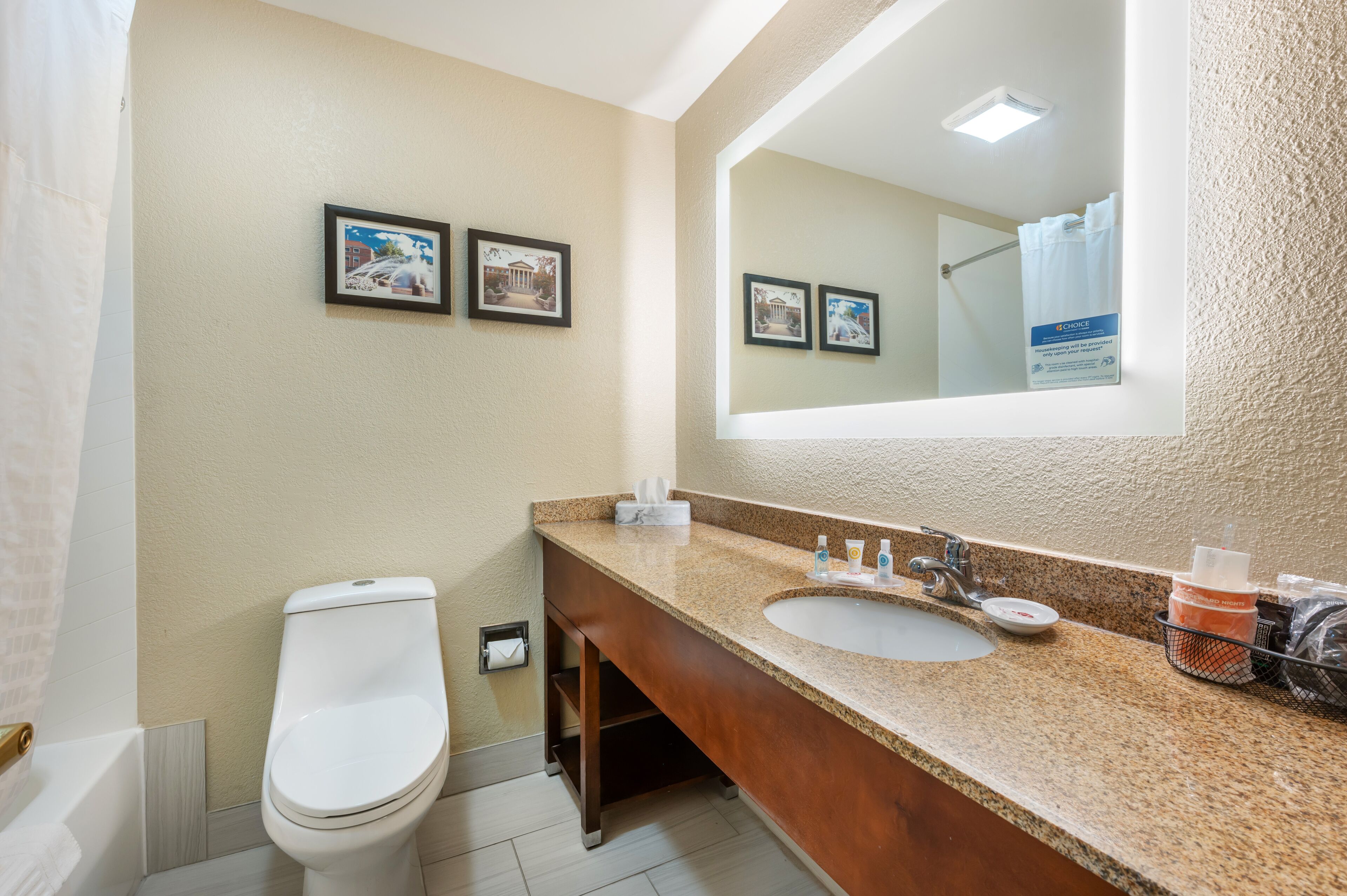 A bathroom with a white toilet, a sink with a granite countertop, and a large mirror. There are two framed pictures on the wall above the mirror.