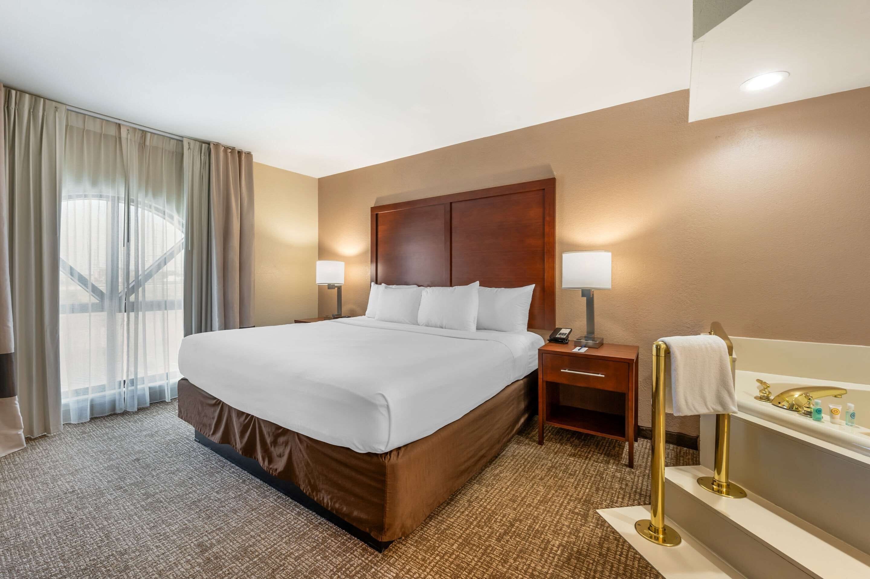 A hotel room with a large bed, a nightstand with a lamp, a bathroom with a bathtub, and a window with curtains.