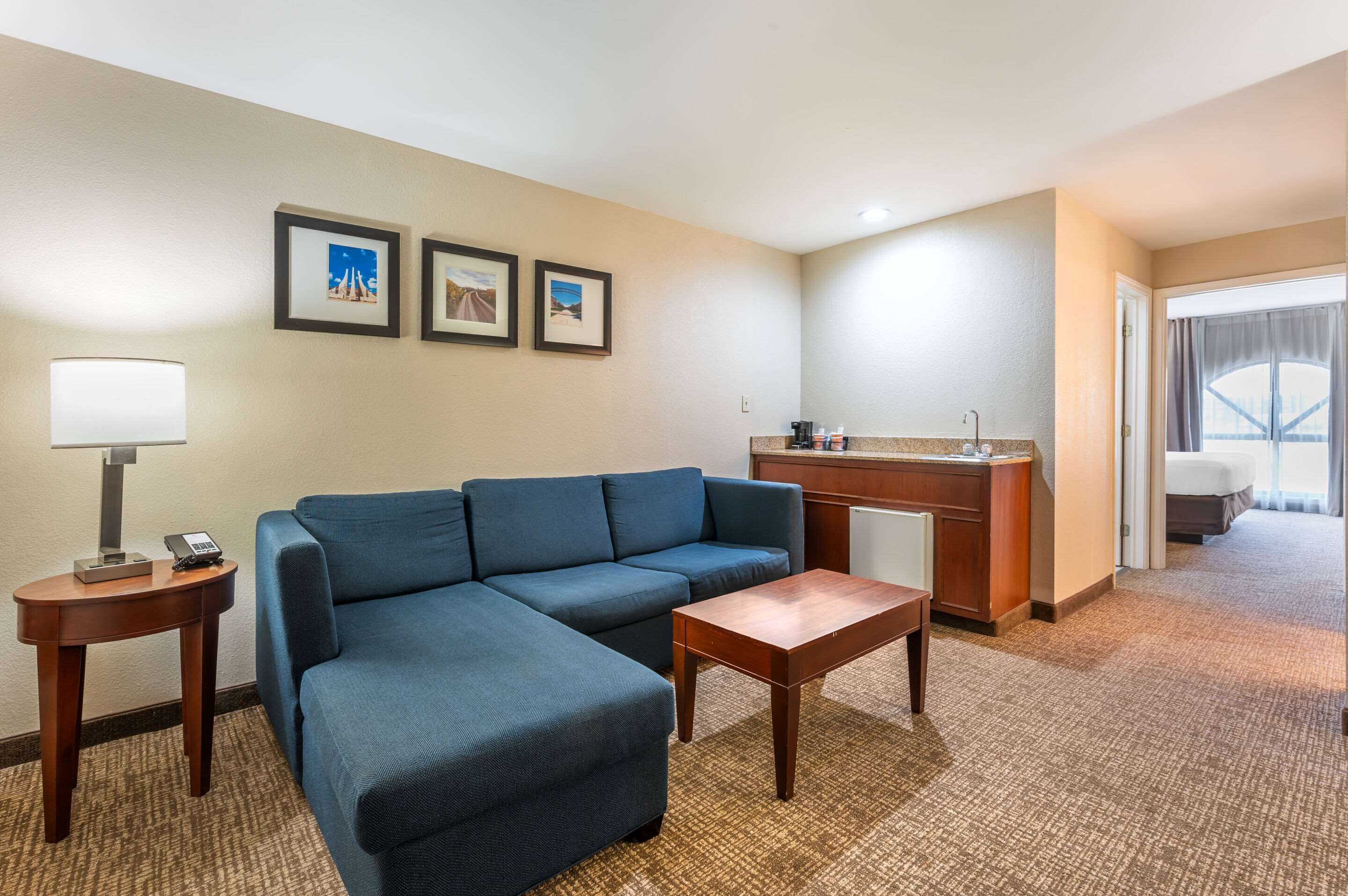 A hotel room with a blue sofa, a small wooden table, a lamp, a minibar, and framed pictures on the wall.