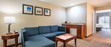 A hotel room with a blue sofa, a small wooden table, a lamp, a minibar, and framed pictures on the wall.