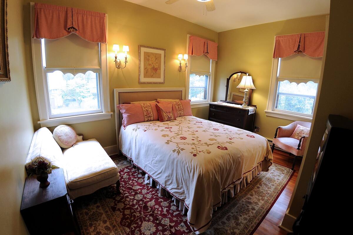 A bedroom with a large bed, a nightstand, a dresser, a chair, and a window with curtains.