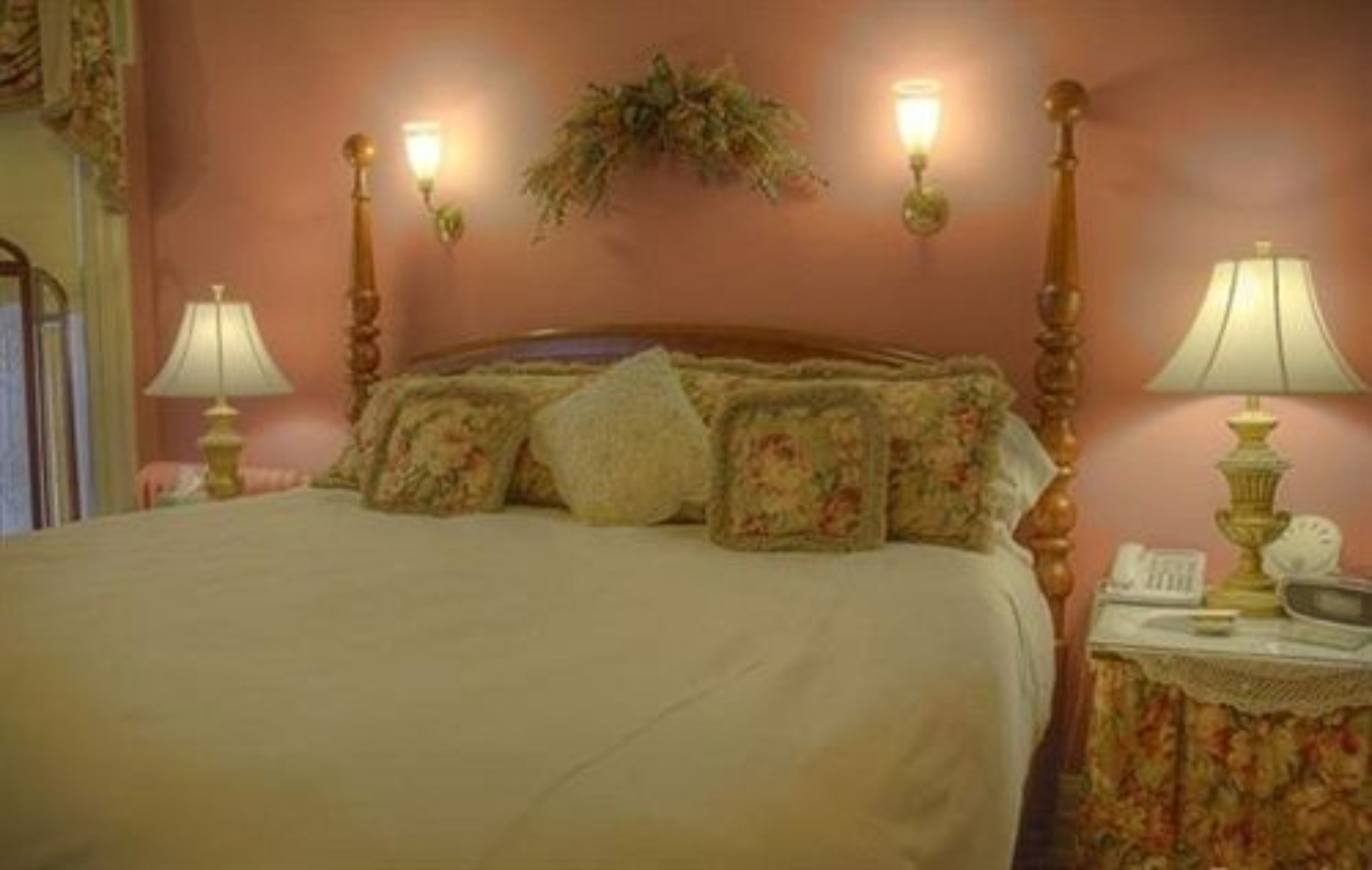 Een slaapkamer met een groot bed, kussens met een bloemenprint, een nachtkastje met een lamp en wandlampen.